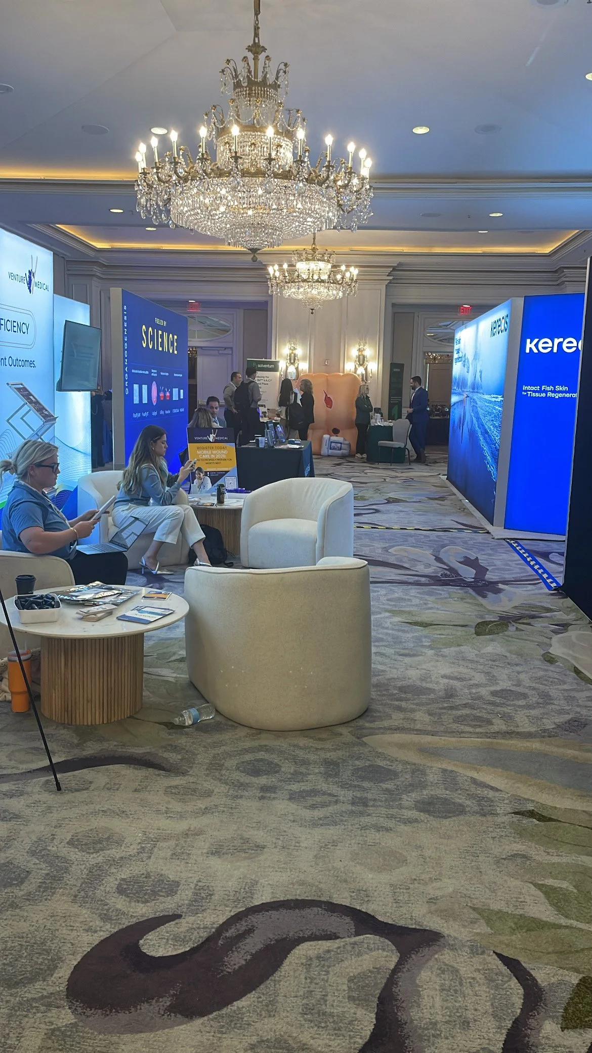 A conference or trade show in an elegant ballroom with chandeliers, displaying booths and attendees engaging with exhibits.