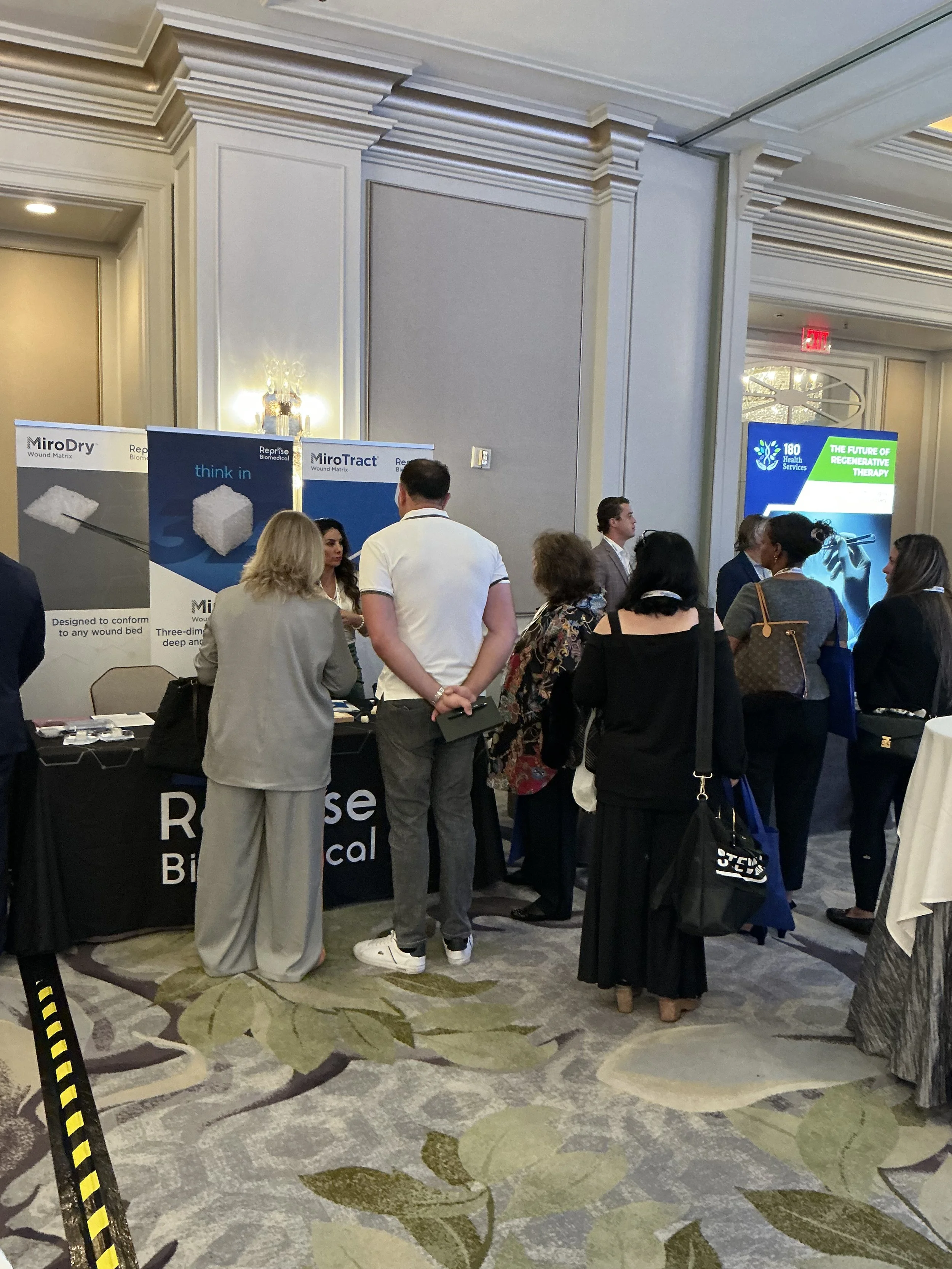 People gathered around exhibition booths at a conference or trade show in a hotel ballroom.