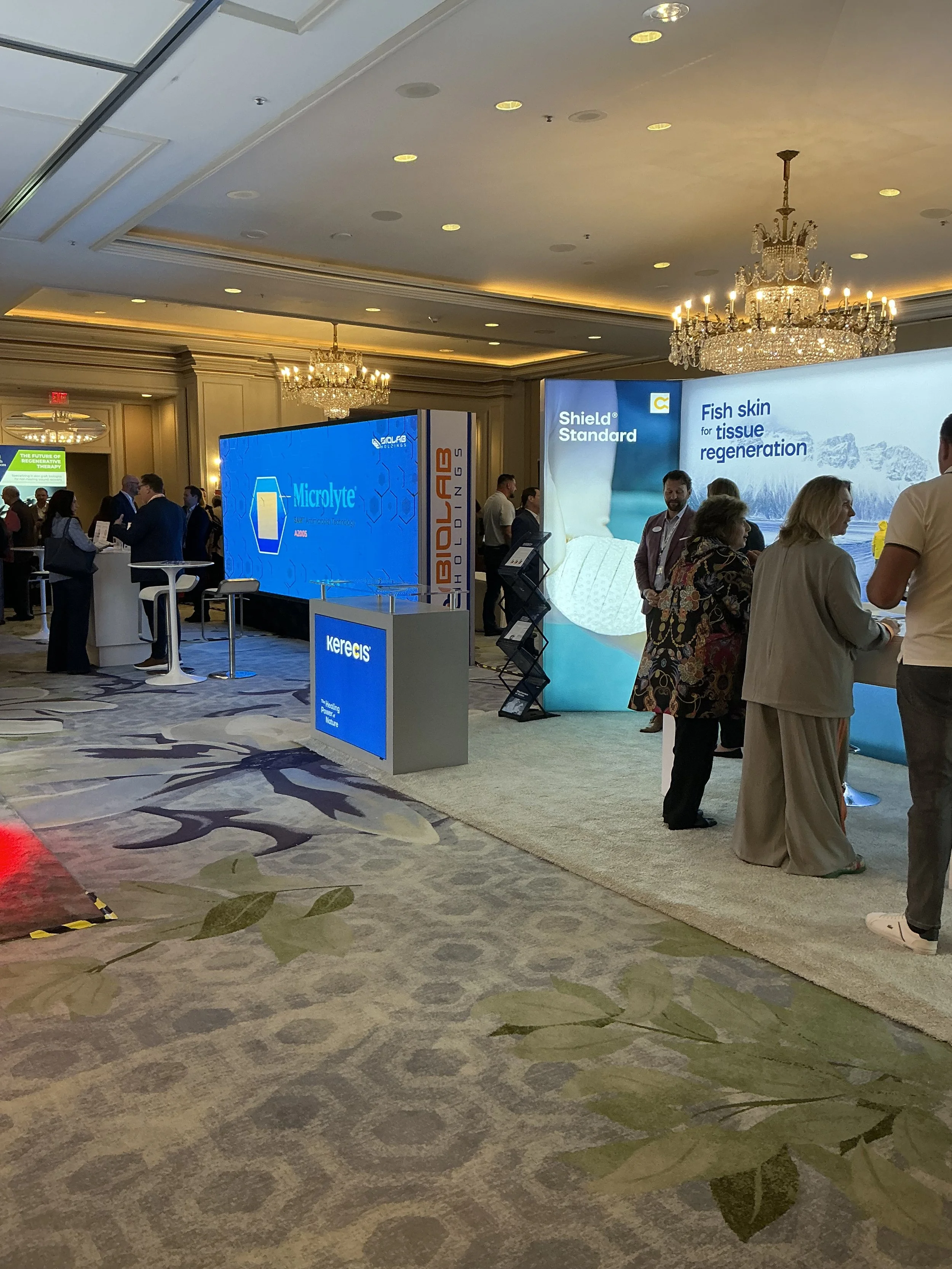 A conference or trade show with various booths and people engaging with displays. Prominent screens display information about medical or skincare products, including one with text about fish skin tissue regeneration. Elegant chandeliers hang from the