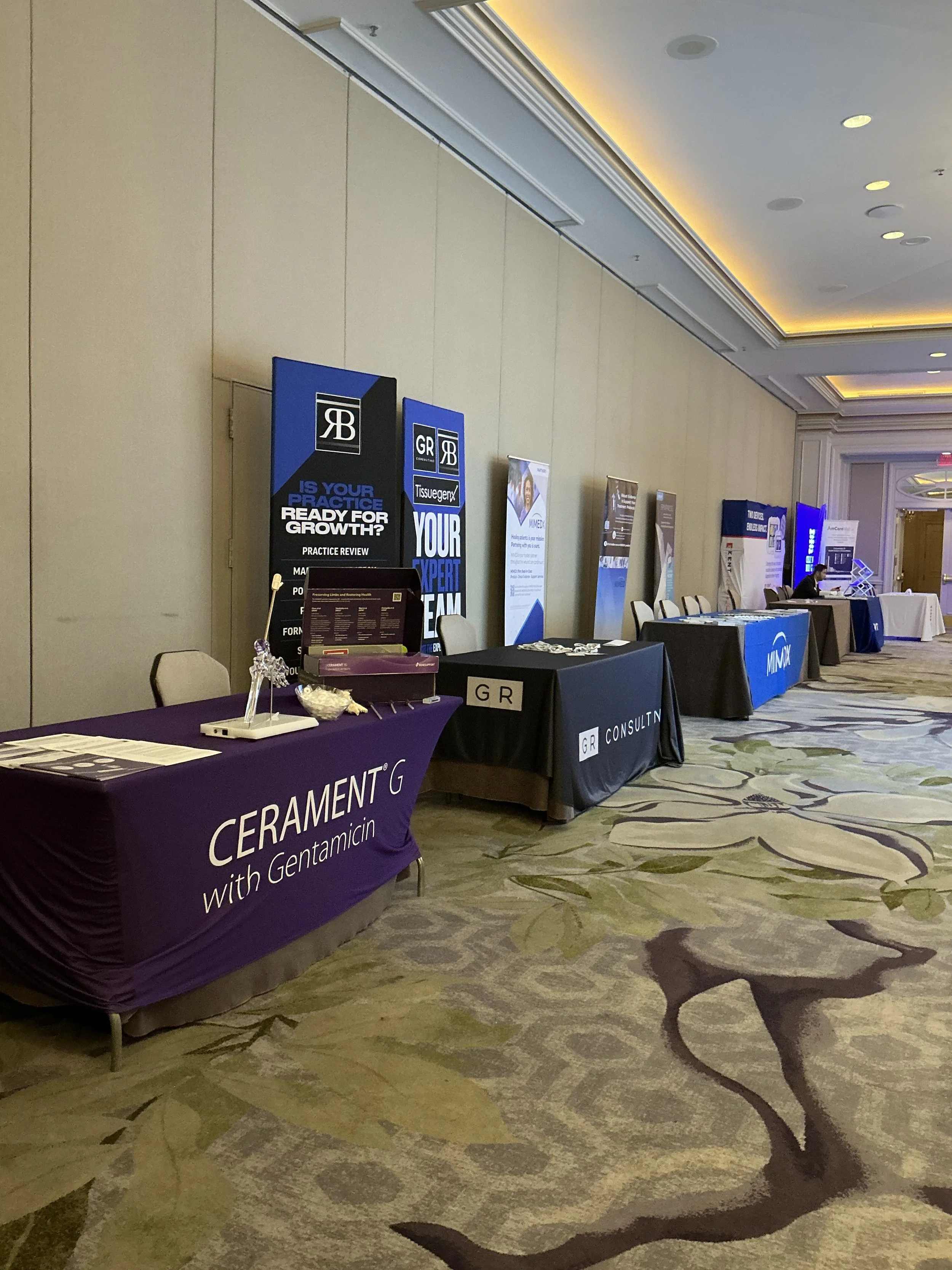 Photo of an exhibition hall with several booths set up with banners and tables, some with promotional materials, in a hotel conference room.