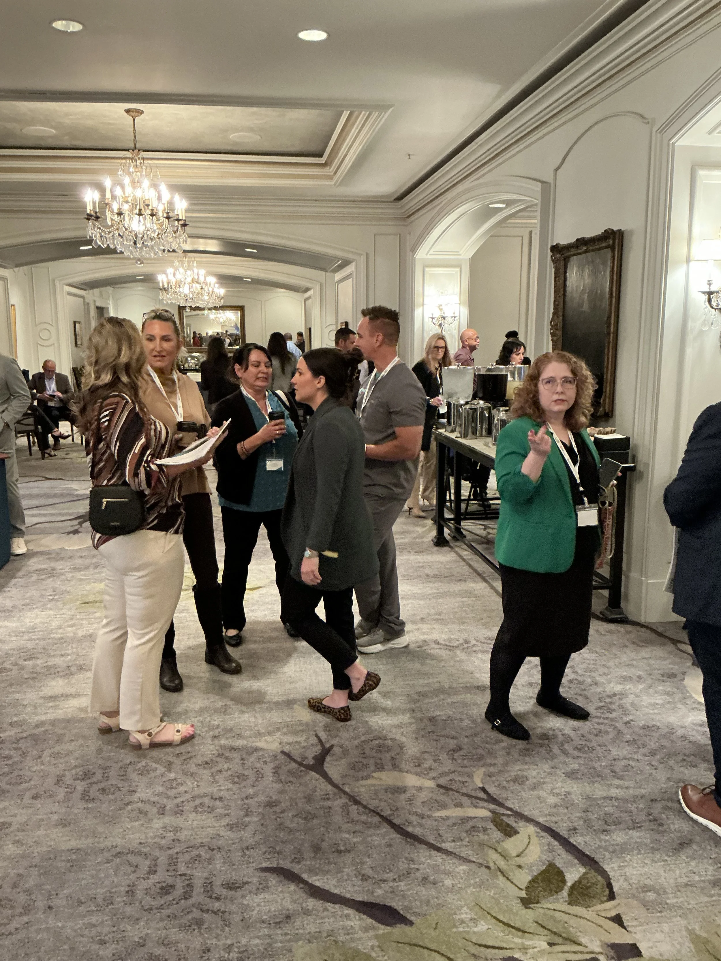 People attending a conference or event in a luxurious hotel ballroom with chandeliers, ornate wall moldings, and a refreshment table.