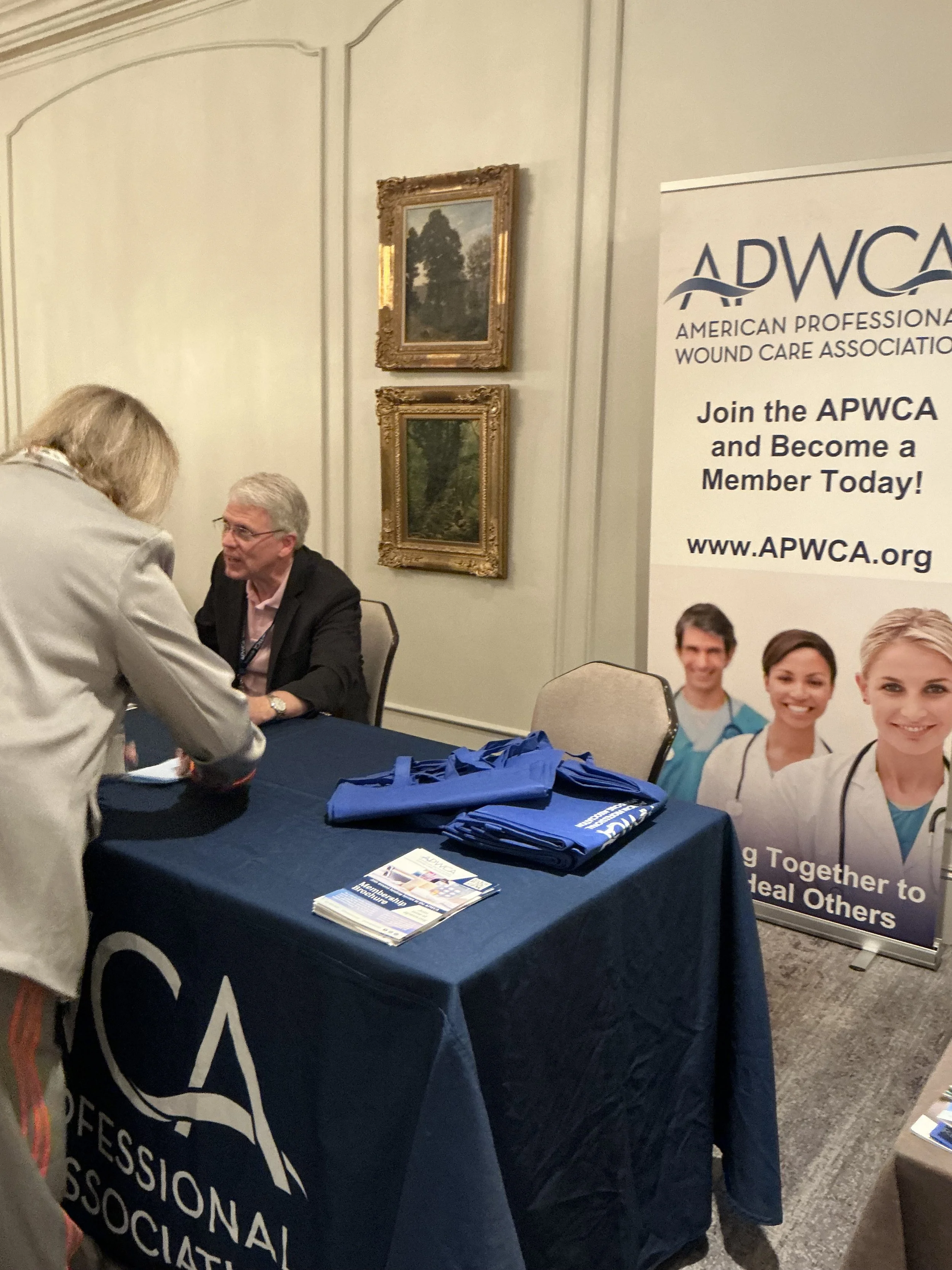 A booth at a conference for the American Professional Wound Care Association (APWCA), with a table covered by a dark blue tablecloth displaying brochures and tote bags, a standing banner with images of healthcare professionals, and two people engaged