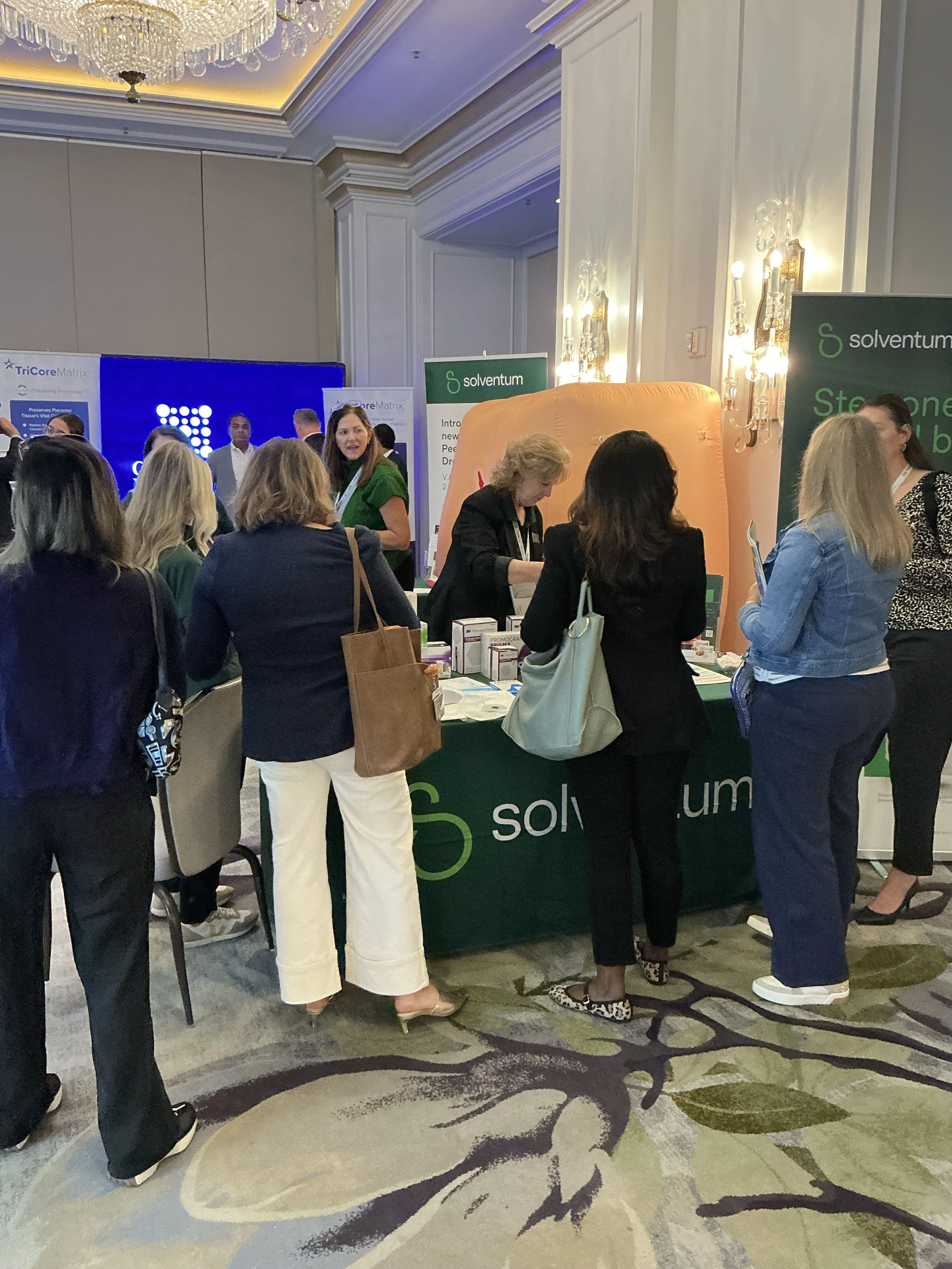 Group of people gathered around a booth at a conference, with banners displaying 'solventum' and 'TriCore Matrix,' in a ballroom with a chandelier and decorative wall lighting.