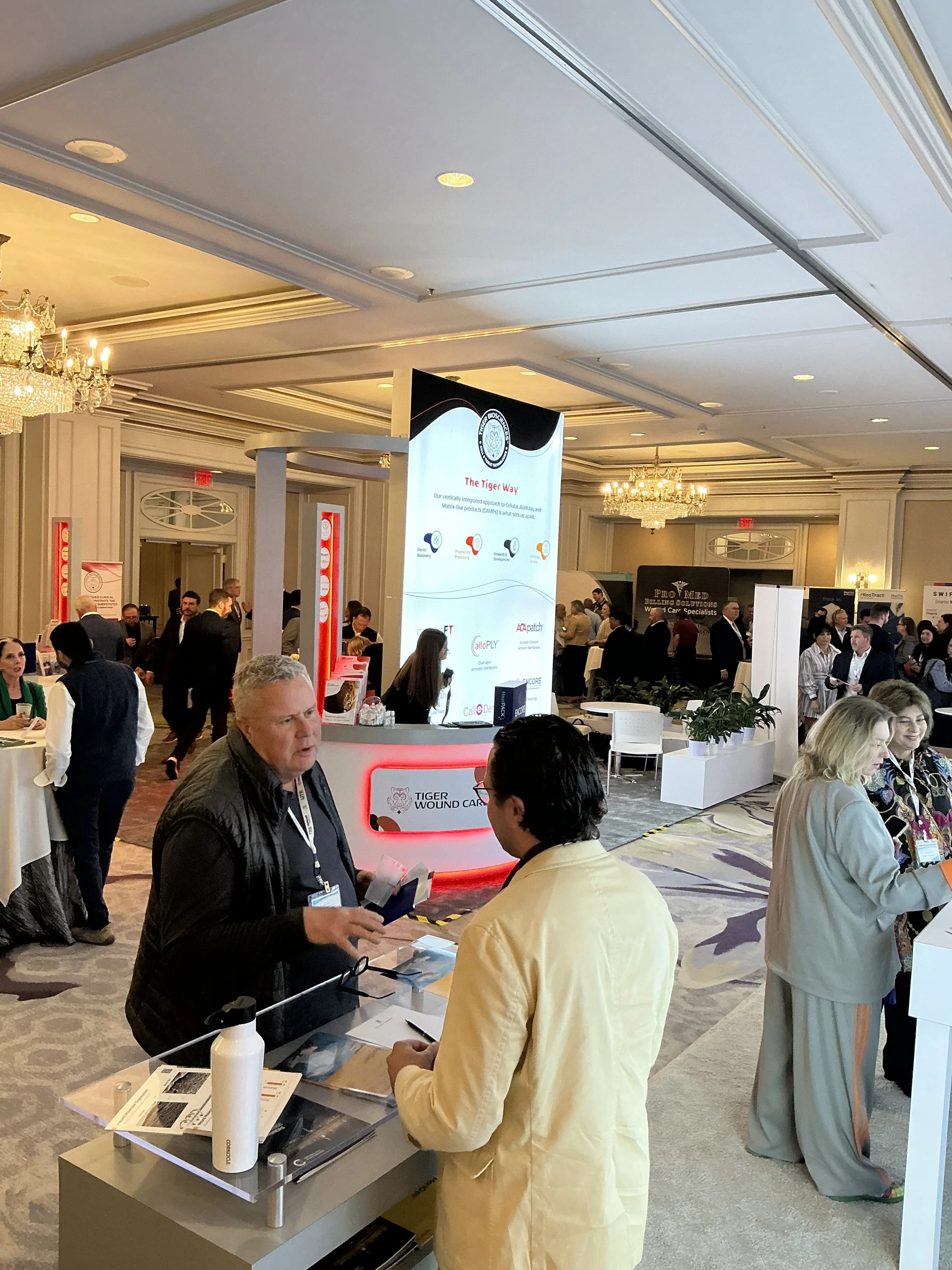 People attending a conference or trade show in a spacious, well-lit ballroom with ornate chandeliers. A large digital screen displays a presentation, and several booths and tables are set up throughout the room. Attendees are engaging with exhibitors