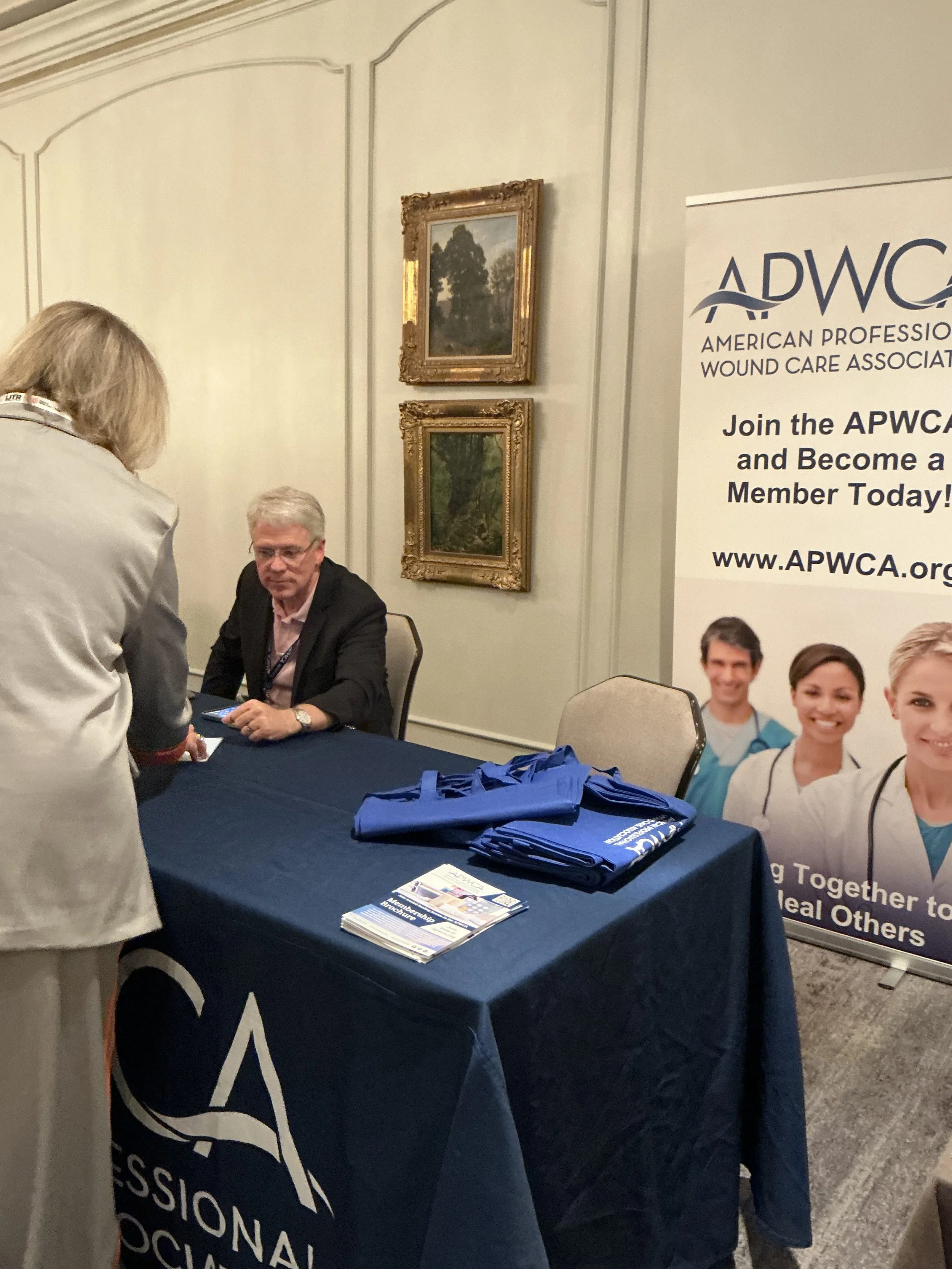 A person in a gray suit talks with a man sitting behind a table at a conference or event. The table has flyers and blue tote bags. A banner for the American Professional Wound Care Association (APWCA) stands beside the table, displaying images of div