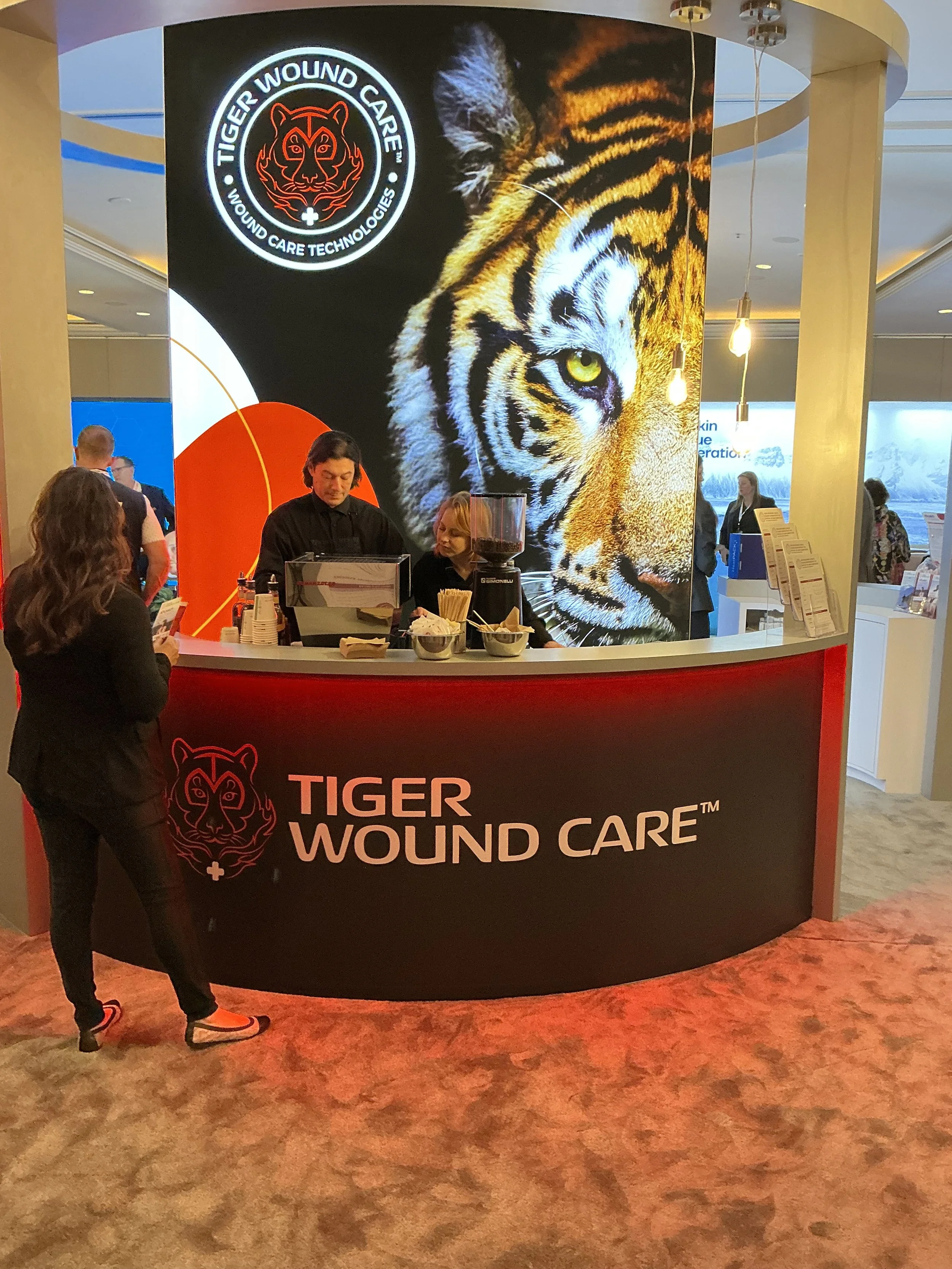 A booth at an event promoting Tiger Wound Care features a large background screen displaying a close-up image of a tiger's face and the company logo. The booth has a curved black counter with the company name and logo. Two staff members are behind th