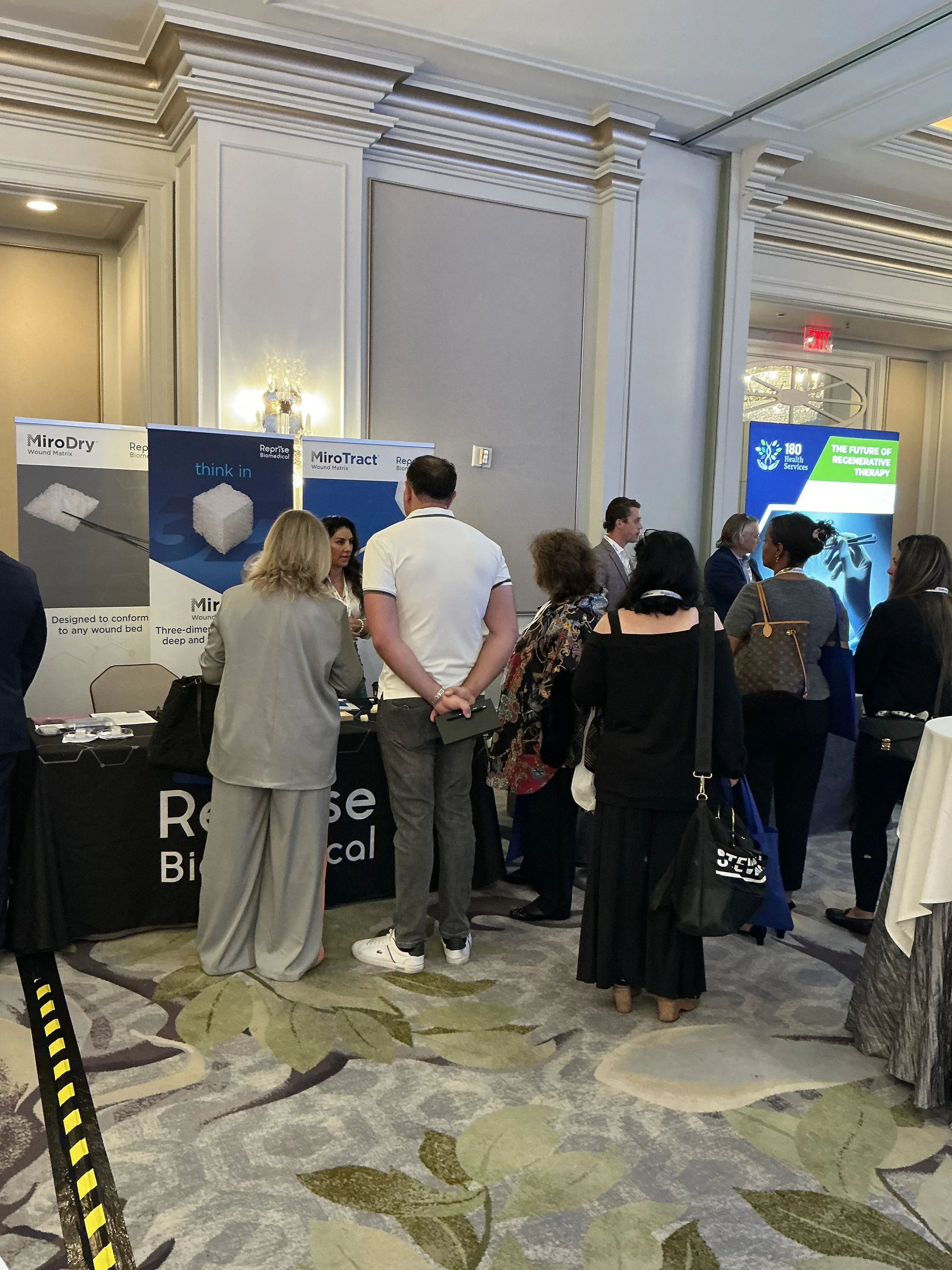 A group of people gathered around a medical booth at a conference or trade show. The booth displays banners for Repose Medical and has a table with promotional materials. In the background, a digital screen advertises regenerative therapy, and the se