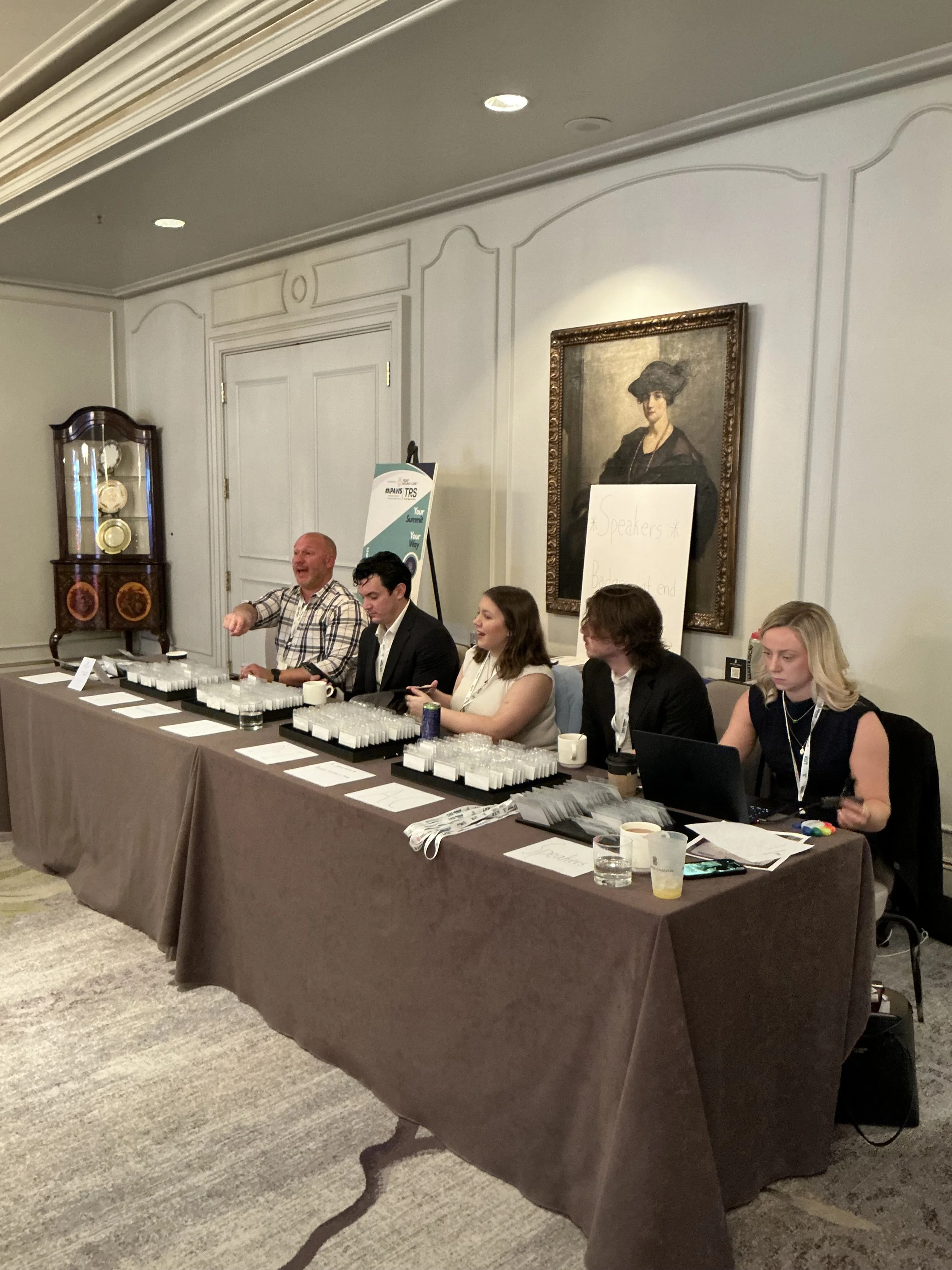 A panel of five people sitting at a long table during a conference or meeting, with name tags, water glasses, and notebooks in front of them. There is a framed portrait on the wall behind them and a sign that says 'Speakers'.