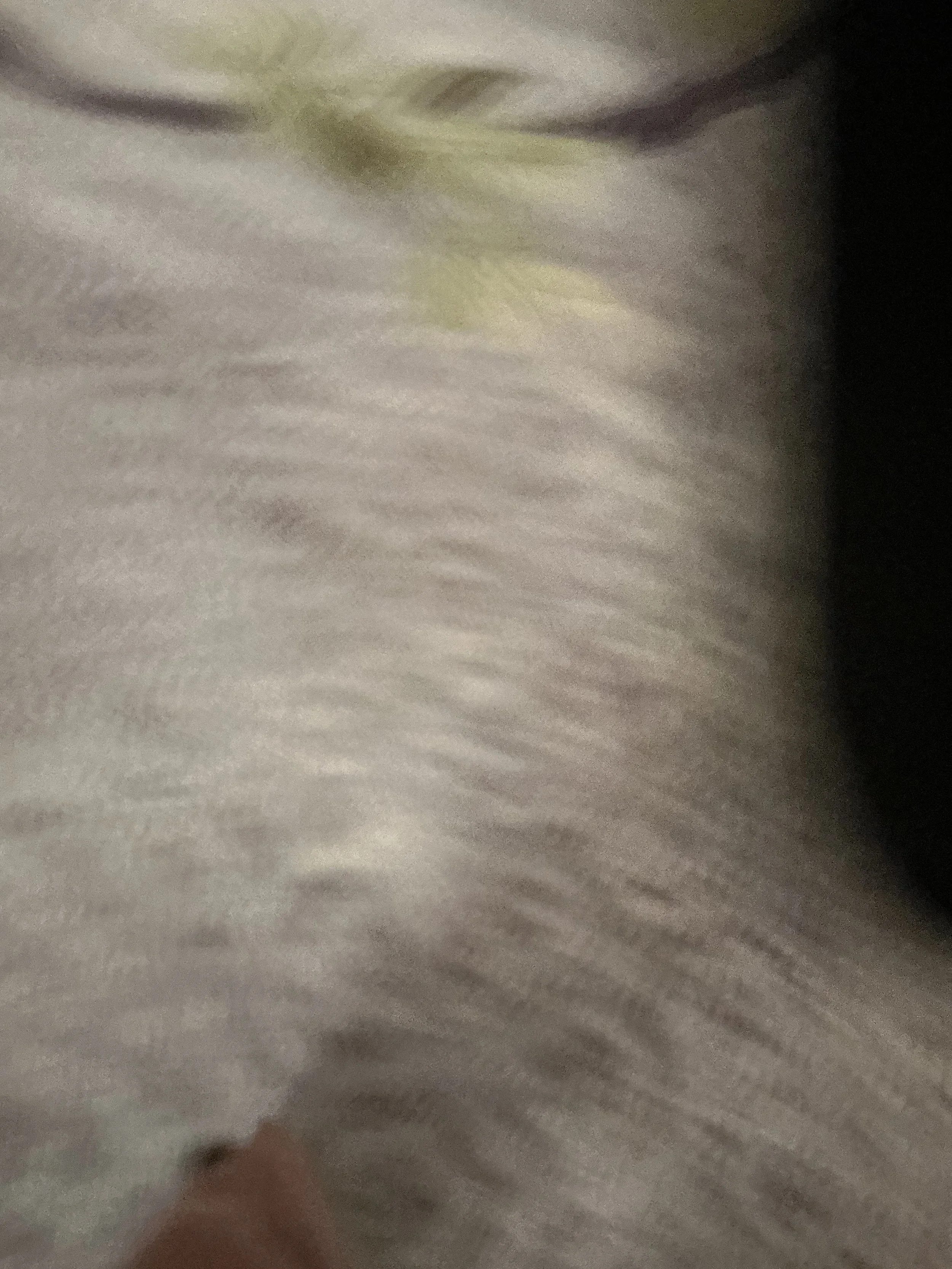 Close-up of gray and yellowish fabric, possibly a blanket or pillow, with a blurred hand or finger at the bottom.