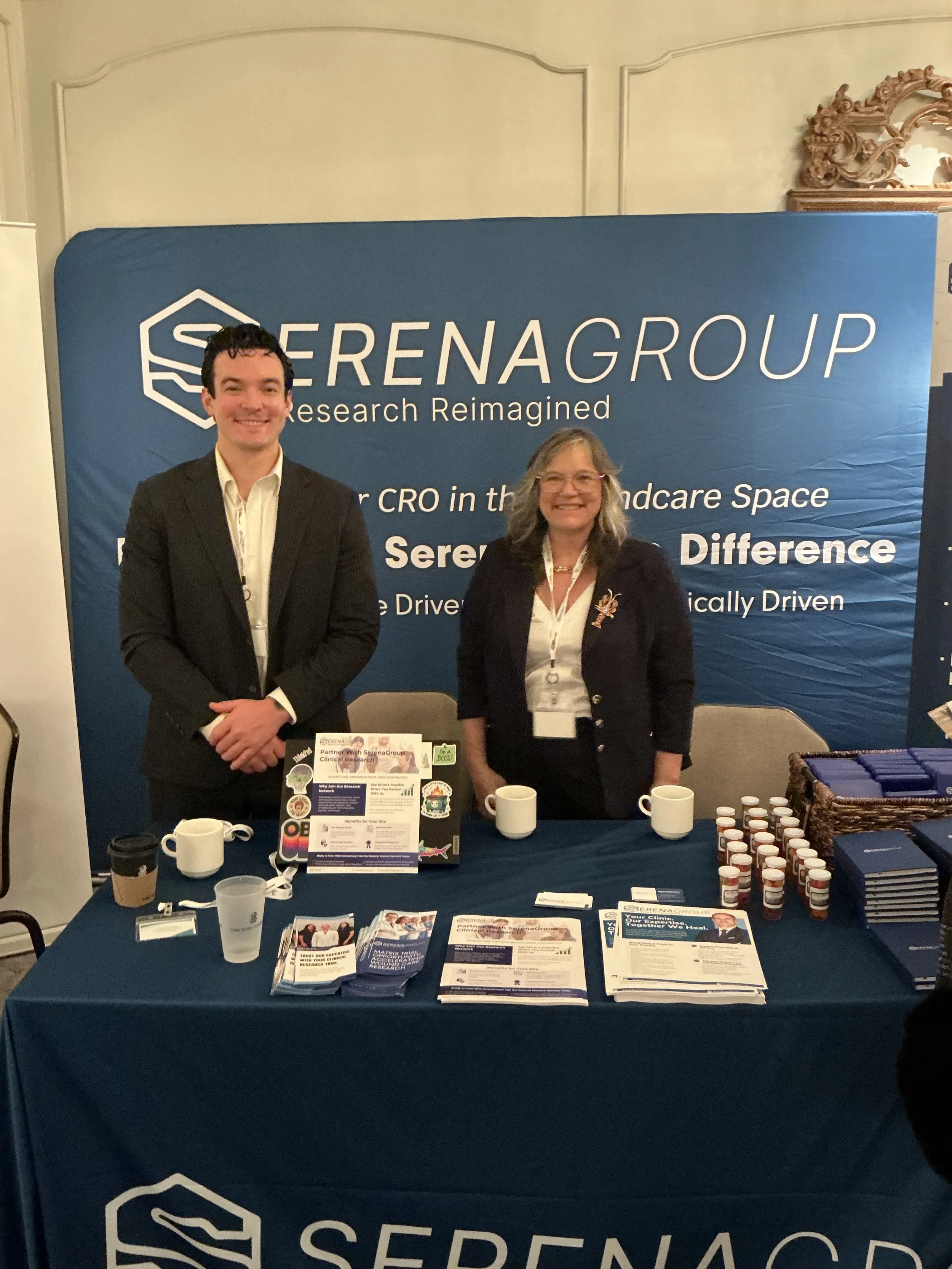 Two people standing behind a table at a conference booth with a blue backdrop that says 'SERENAGROUP, Research Reimagined' and 'Partner with Serenagroup,' with promotional materials, brochures, and small bottles of medicine or supplements on the tabl