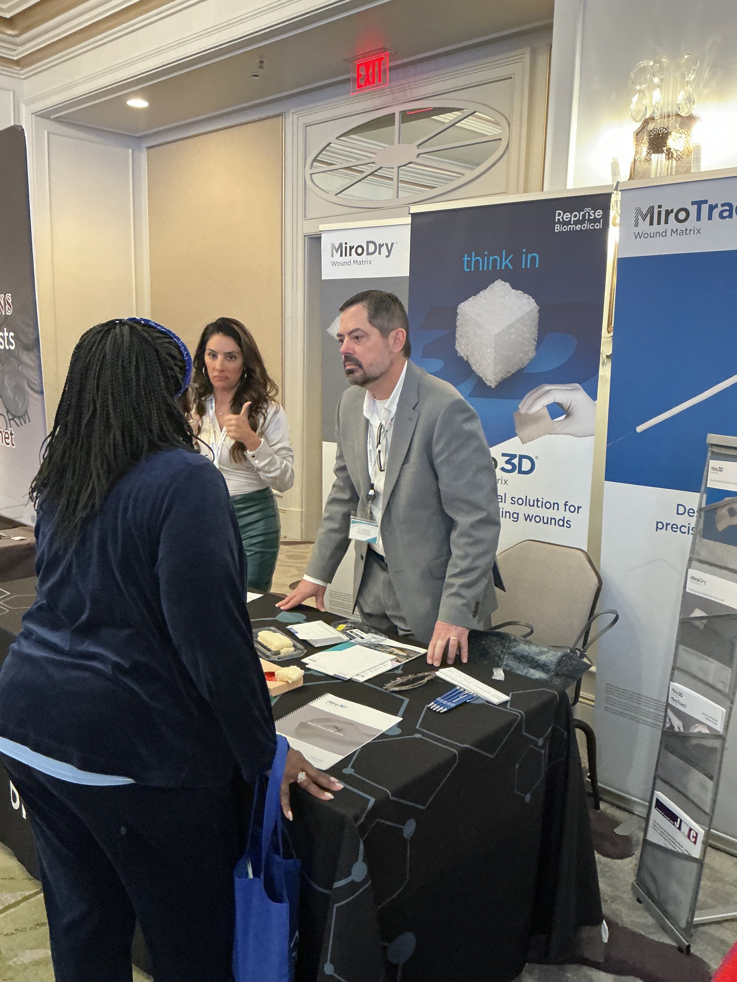 A man in a gray suit and a woman with dreadlocks having a conversation at an exhibition booth about wound treatment products. The booth features banners promoting MiroDry and MiroTrace wound matrices, and has informational brochures and samples on th