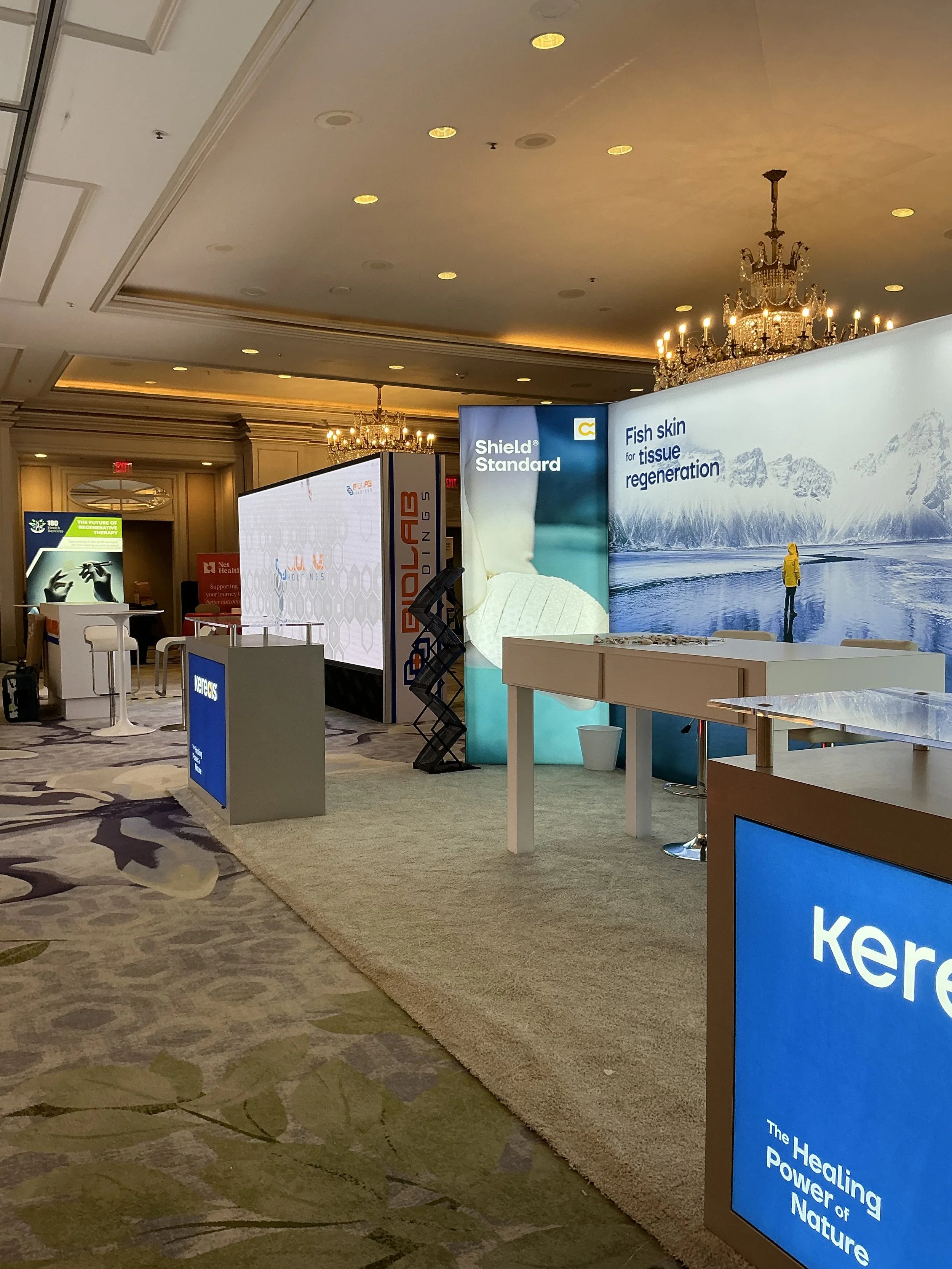 Exhibition booth with digital displays and informational signage about skincare and regenerative medicine in a conference room, featuring chandeliers and carpeted flooring.