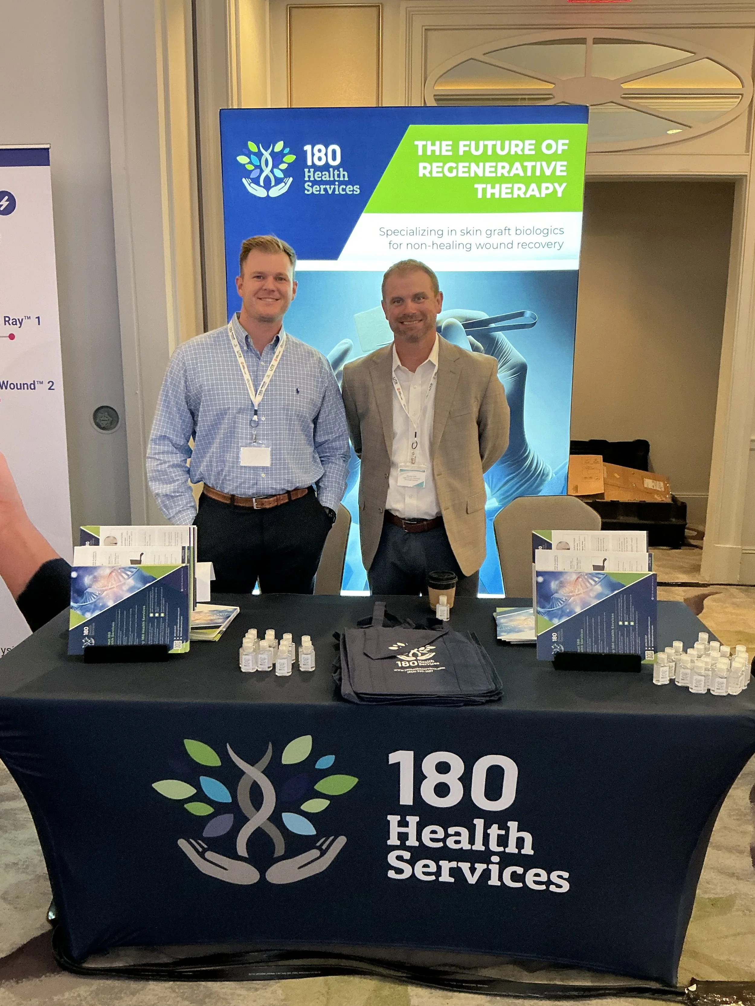 Two men standing behind a promotional table for 180 Health Services, with a large banner that reads 'The Future of Regenerative Therapy.' The table displays informational pamphlets, small bottles, and a tote bag with the company logo.