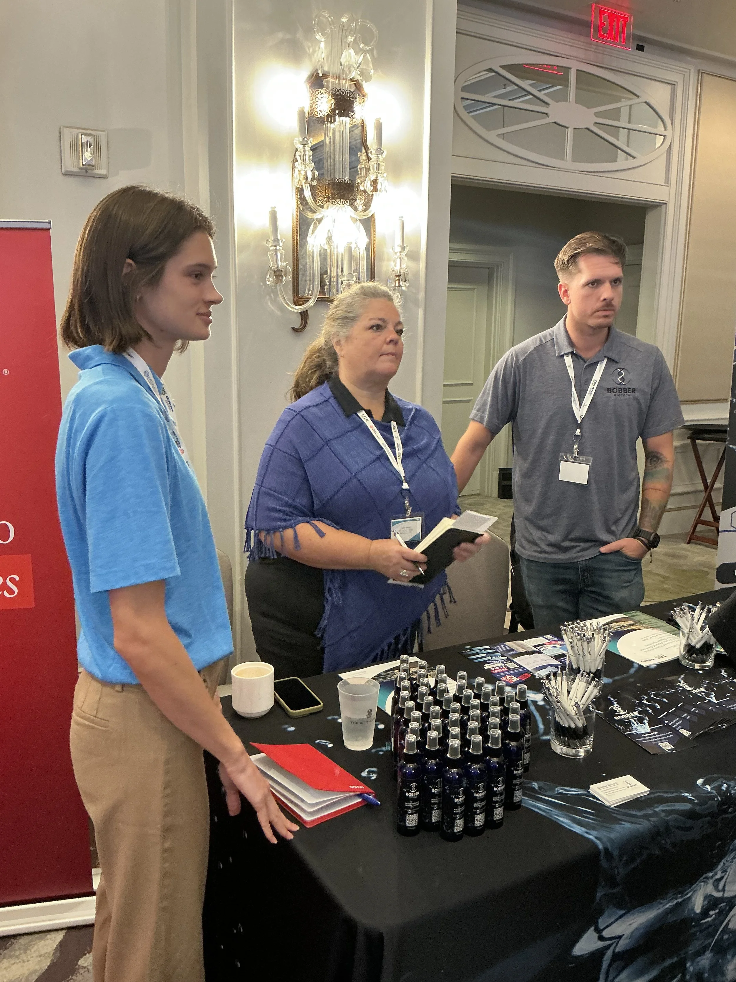 Three people standing behind a table with promotional items and pens, at a conference or expo, with a chandelier and an exit sign in the background.
