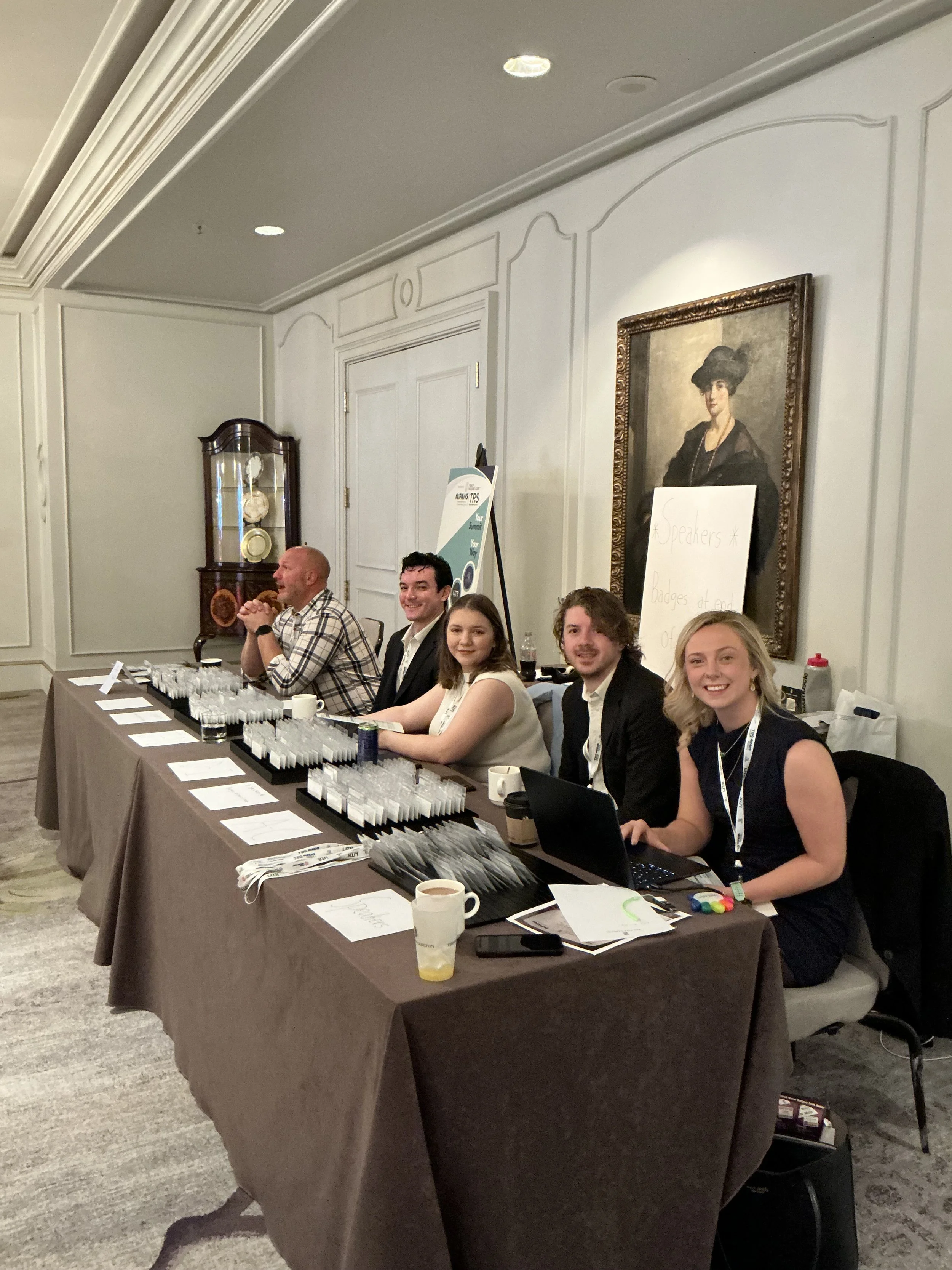 People sitting at a registration or information table at an event, with papers, pens, and laptops in front of them, in a formal room with framed portrait and a china cabinet.