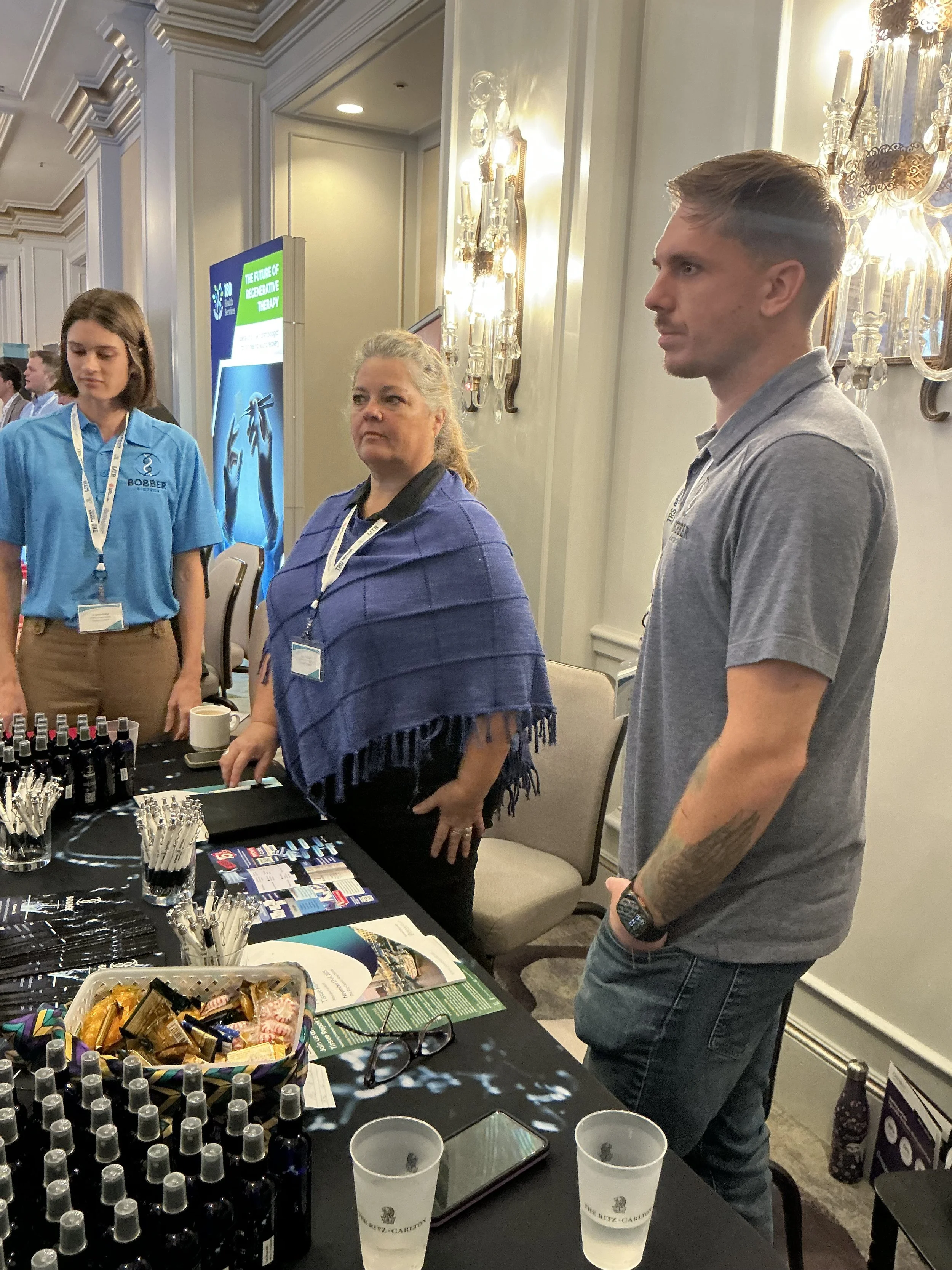 People standing at a booth with bottles, pens, and brochures, in a conference room or event space.