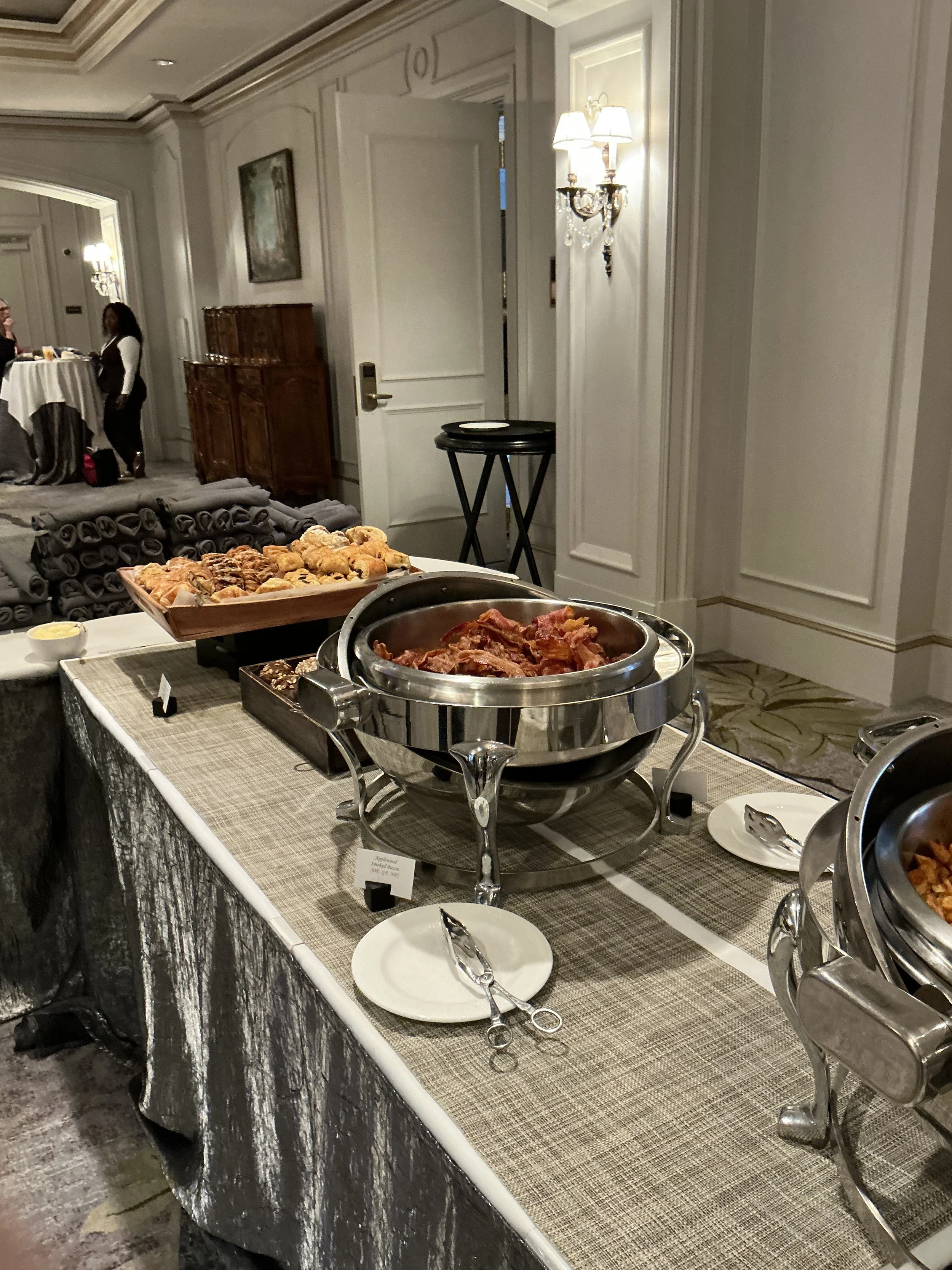 Buffet table with trays of bacon, pastries, and other breakfast items in a hotel or event space, with people in the background.