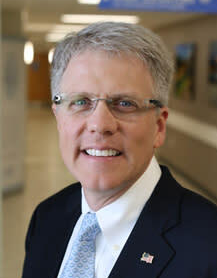 A smiling middle-aged man with gray hair and glasses, wearing a dark suit, white shirt, and a patterned light blue tie, standing indoors with a blurred background.