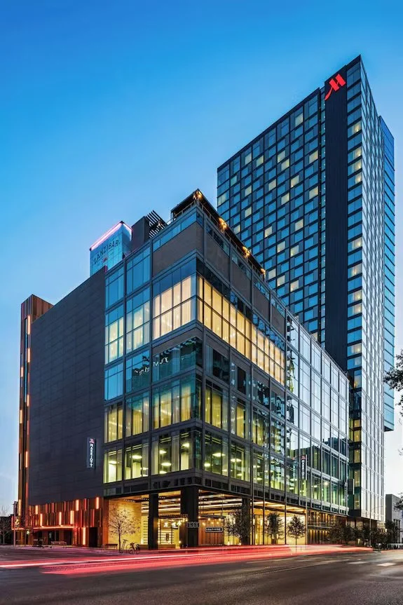 Modern glass office building with illuminated interior, tall skyscraper with a red logo behind, and a blue evening sky.