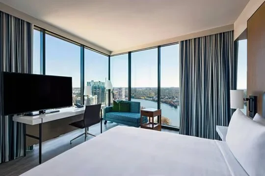 A hotel room with a large bed, a TV on a desk, a green sofa, side table, and floor-to-ceiling windows with city and water views.