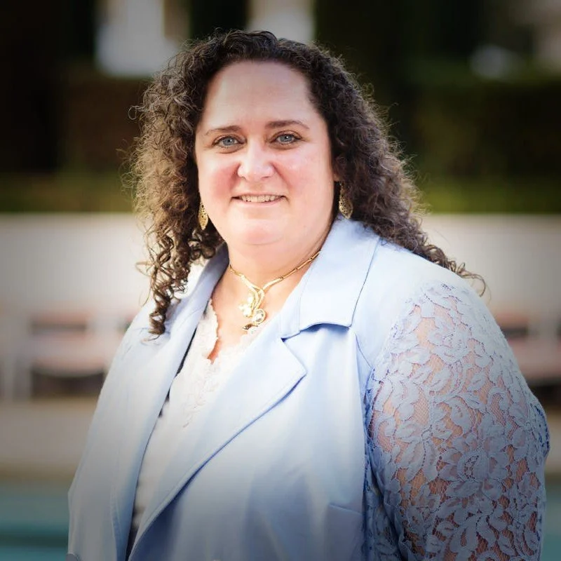 A woman with curly brown hair and blue eyes smiling outdoors, wearing a light blue blazer with lace sleeves and gold jewelry.