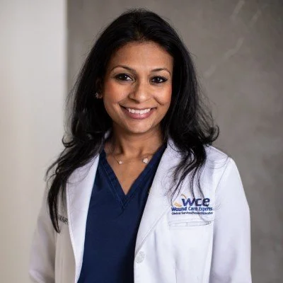 A smiling woman with dark hair wearing a white medical coat and navy scrubs, standing indoors.
