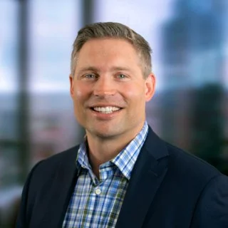 A professional man in a suit with a blue checkered shirt smiling, standing in front of a blurred office background.
