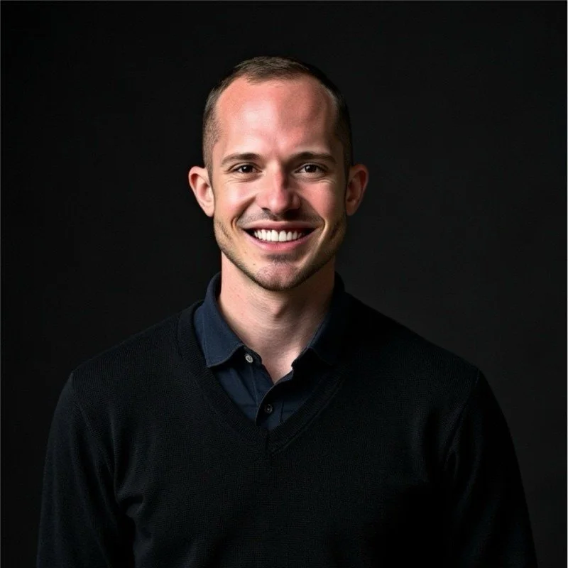 A smiling man with short hair wearing a dark sweater over a collared shirt against a black background.