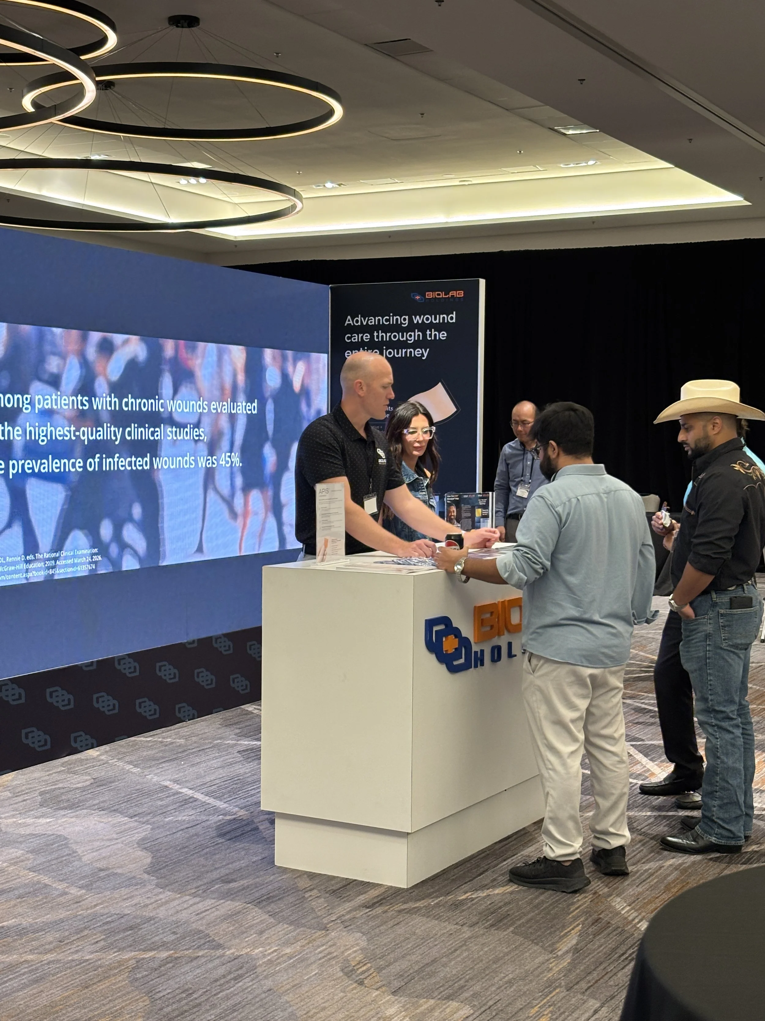 People at a medical conference or expo engaging in conversation at a booth with a large digital screen displaying text about wound care research. The booth has a white counter with branding, and attendees are casually dressed, talking.  TRSS & AWCS
