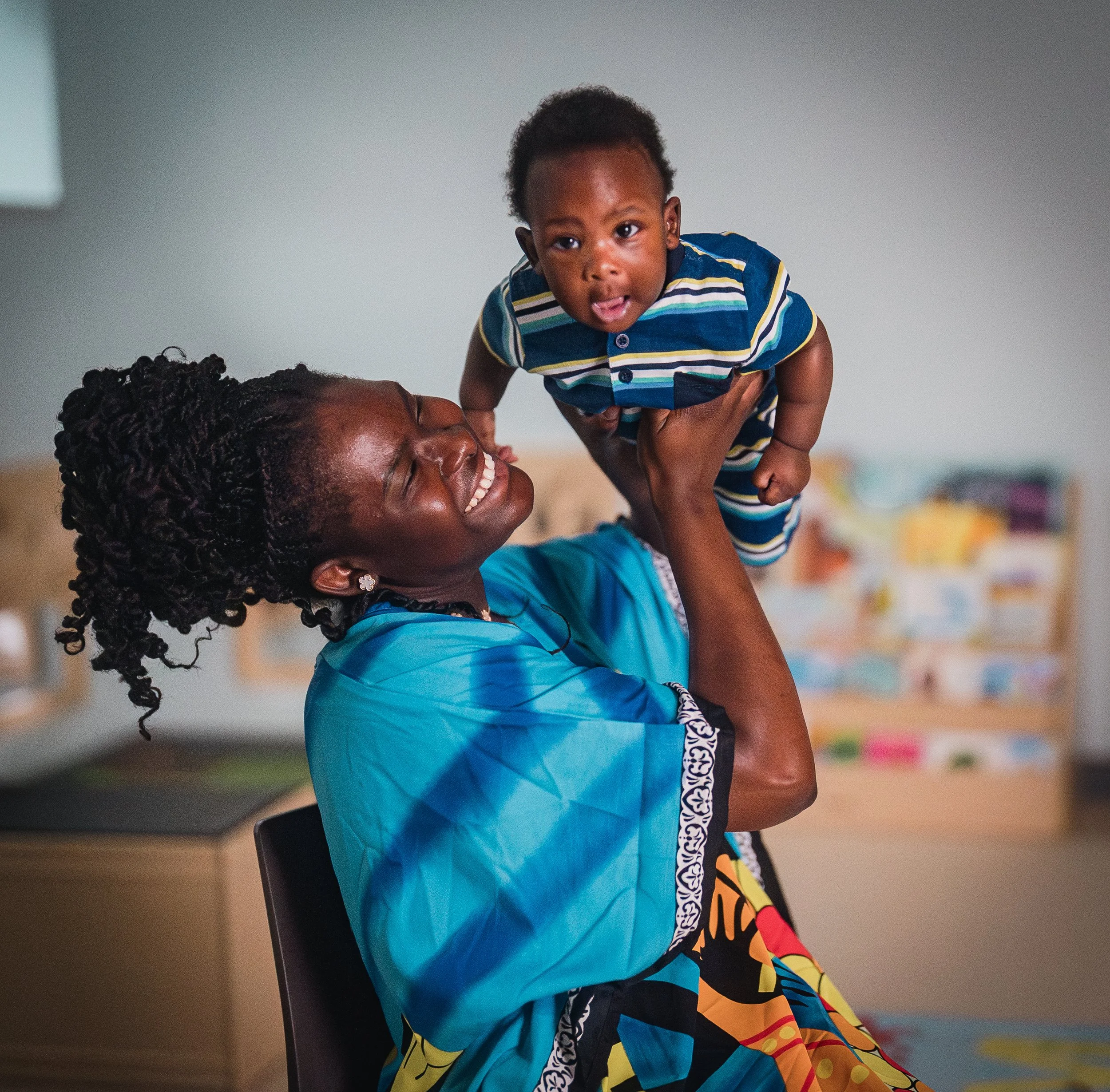 A smiling woman with braided hair holding a young boy in a colorful striped shirt. The boy is smiling with his tongue slightly out, looking directly at the camera. The background is blurred, showing an indoor space with some colorful objects.