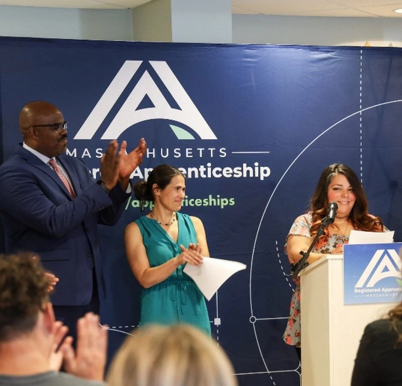 Three people on stage at a Massachusetts apprenticeship event. A man in a suit is clapping, a woman in a teal dress is holding papers, and another woman at a podium is speaking into a microphone. The backdrop features the Massachusetts Apprenticeship logo and branding.