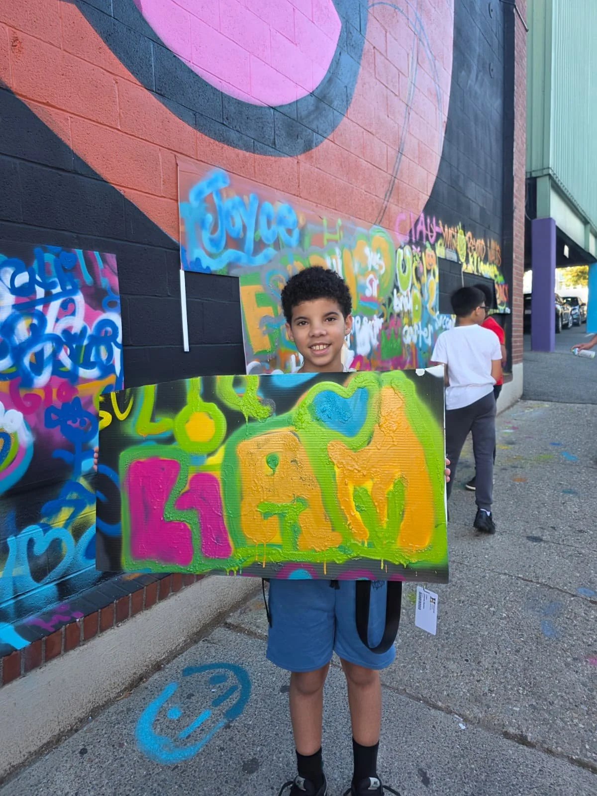Young boy smiling and holding a colorful graffiti-inspired painting in front of a mural wall. Two other boys are visible in the background, also near the mural.