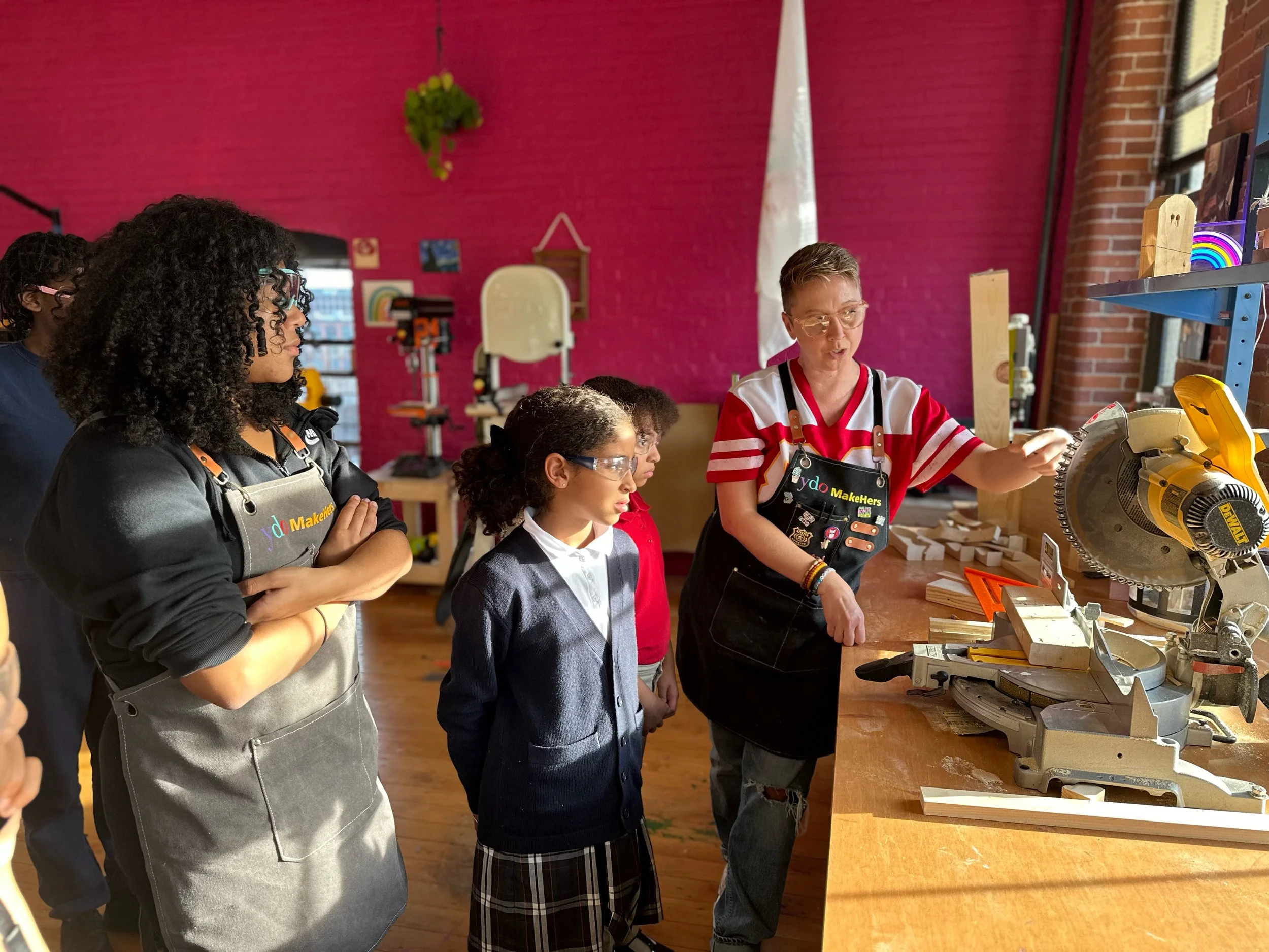 A group of children and an instructor in a woodworking classroom, observing a chop saw. The classroom has a red brick wall and wooden workbenches.