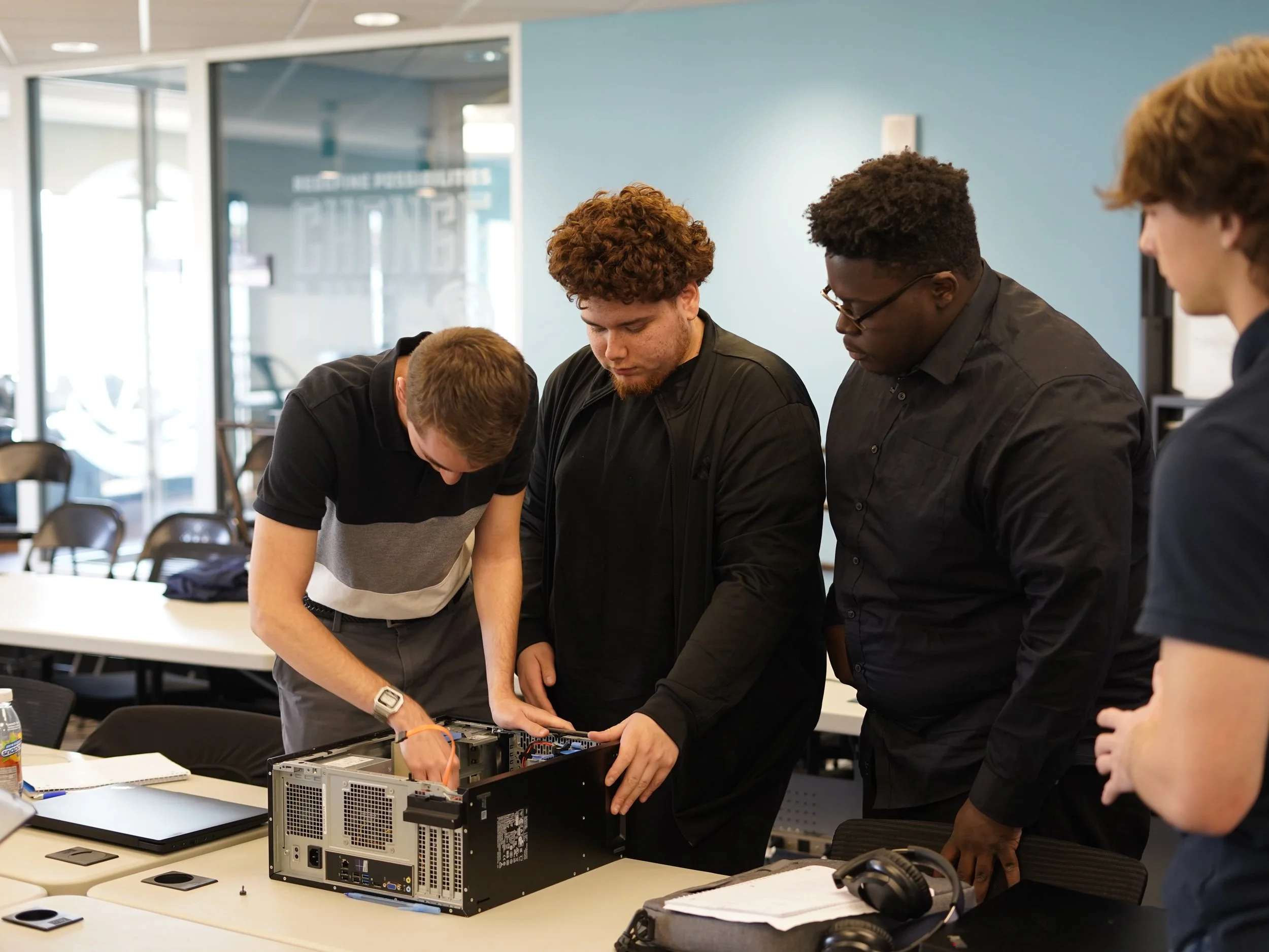 A group of young men working together on a computer inside a modern office or classroom, with tables, chairs, and educational materials visible.
