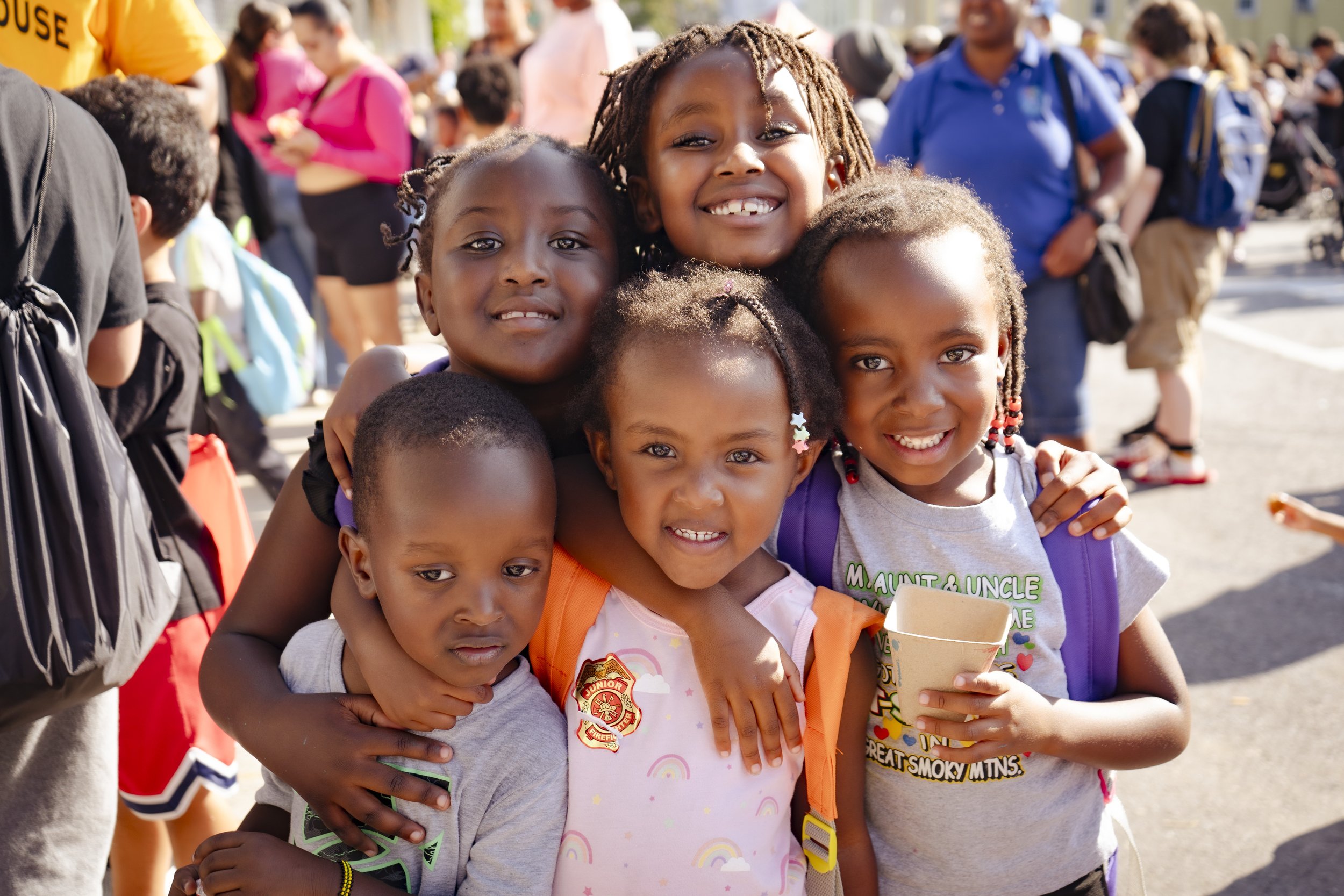 Group of smiling children of various ethnicities embracing at an outdoor event with a crowd in the background.