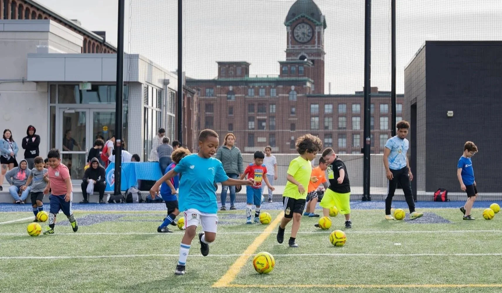 Children participating in a soccer practice on a turf field, with adults and spectators watching in the background, and a cityscape with a clock tower visible behind a protective net.