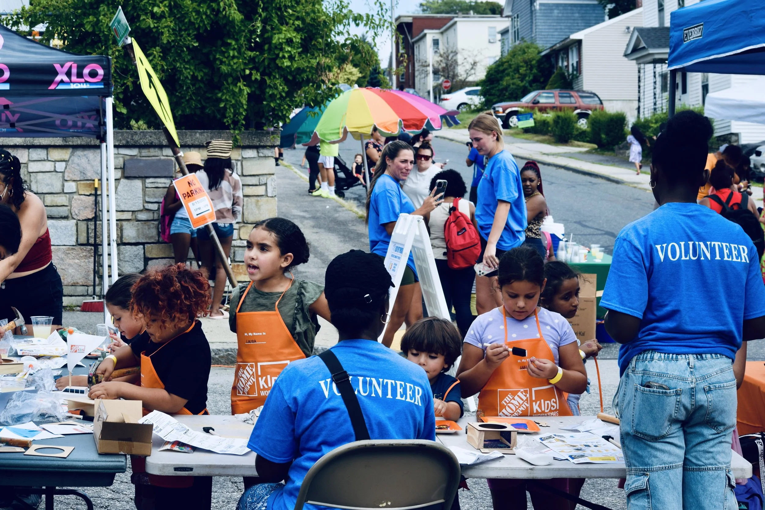 Community event with children and volunteers at outdoor booths, including activities at a table with craft supplies and a colorful umbrella in a residential neighborhood.