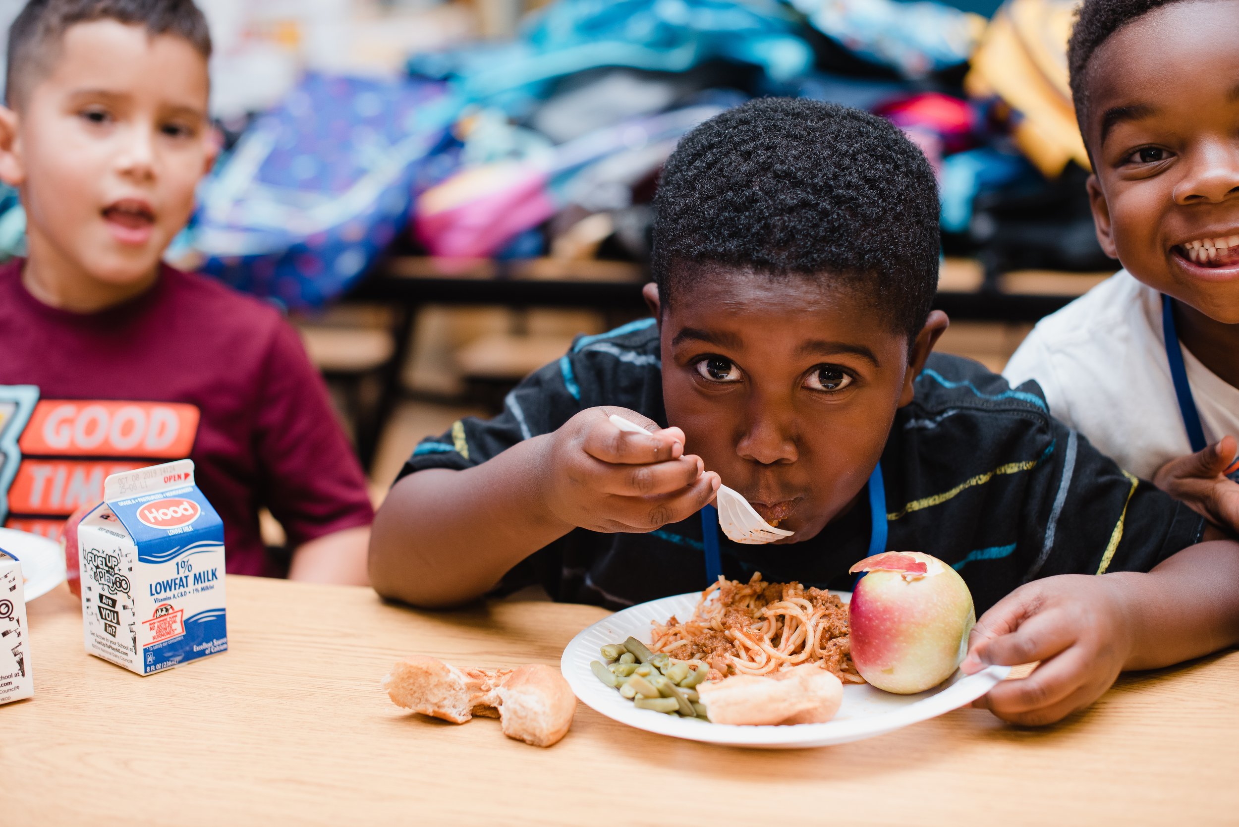 Children eating lunch at a table with food including spaghetti, apple, green beans, bread, and milk.