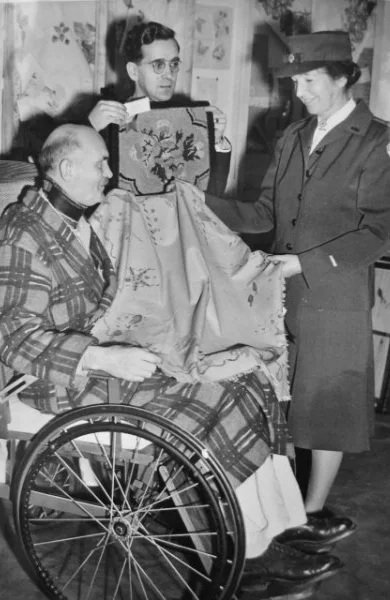 Amelia Peabody in a military uniform and a hat is holding hands with a woman in a wheelchair. Two other women are nearby, one with glasses and short hair, holding a small box, Red Cross.