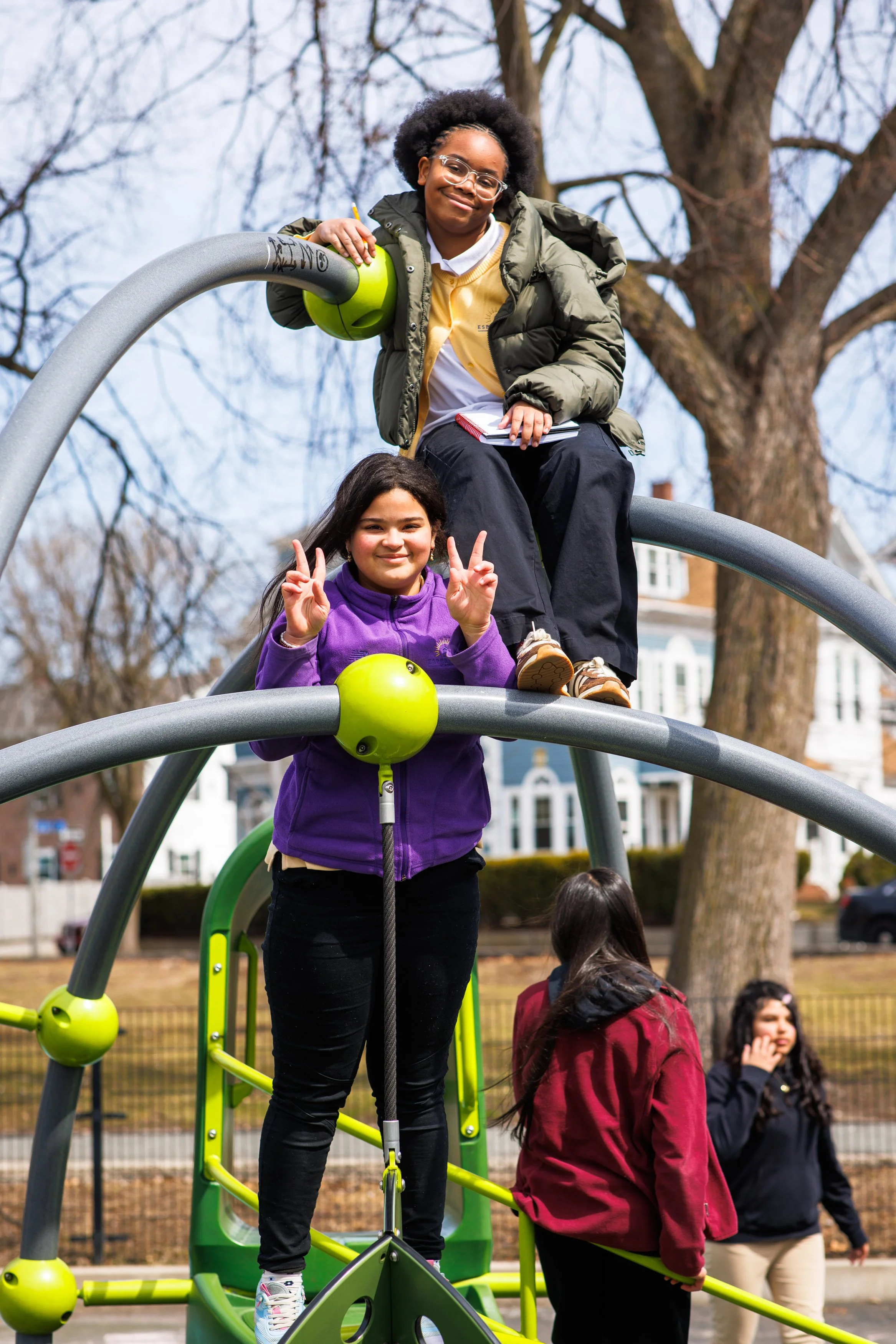 Two young girls play on a green playground structure in a park during daytime. One girl, wearing glasses, a green jacket, and black pants, is sitting on the top level holding a helmet. The other girl, with long dark hair, is standing below, making the peace sign with her fingers. Other children are visible in the background, and leafless trees and houses are also seen.