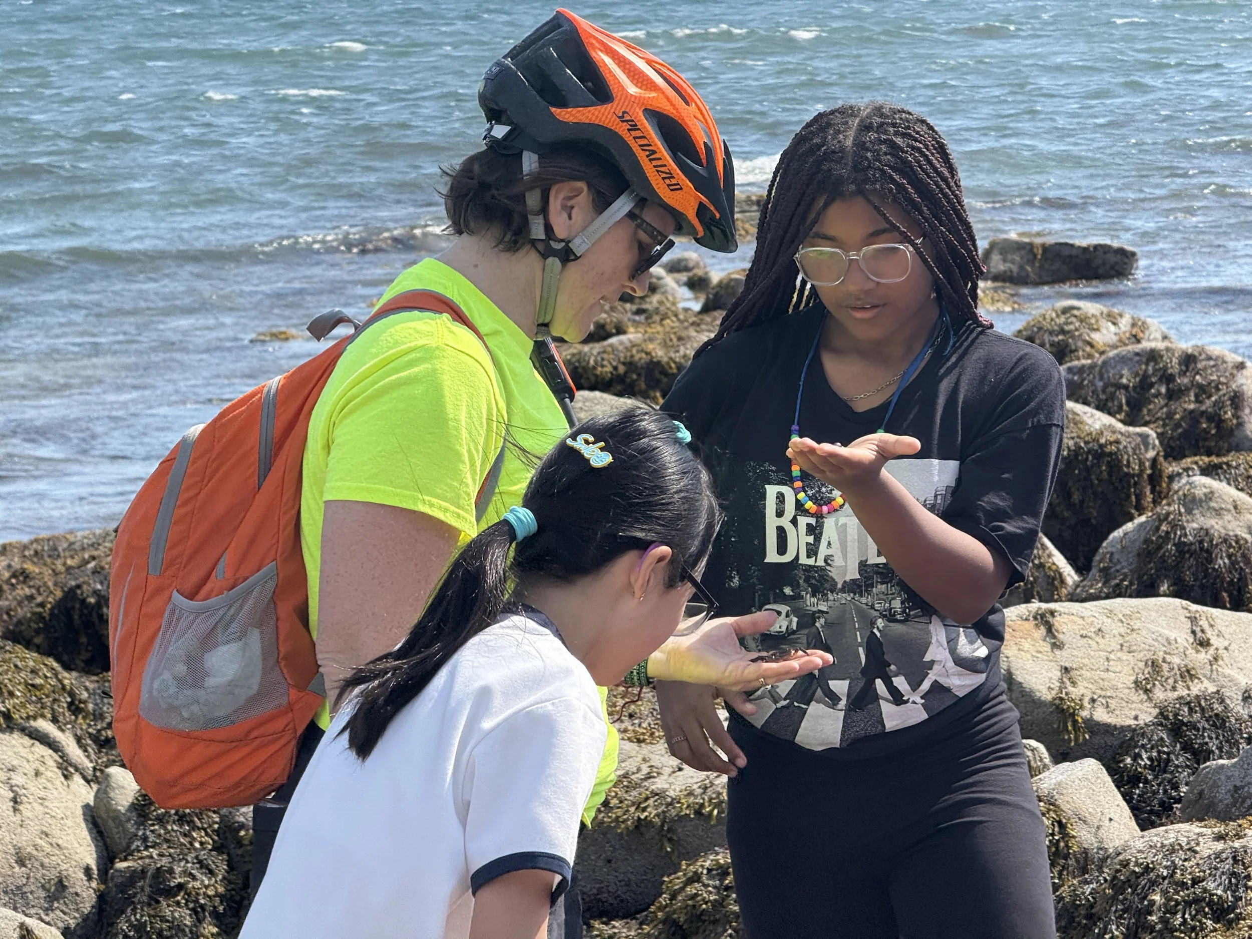 Two young women and a young girl near the rocky shoreline, one woman wearing a helmet and glasses, the girl wearing glasses, and another woman taking a photo or examining something, with ocean waves in the background.