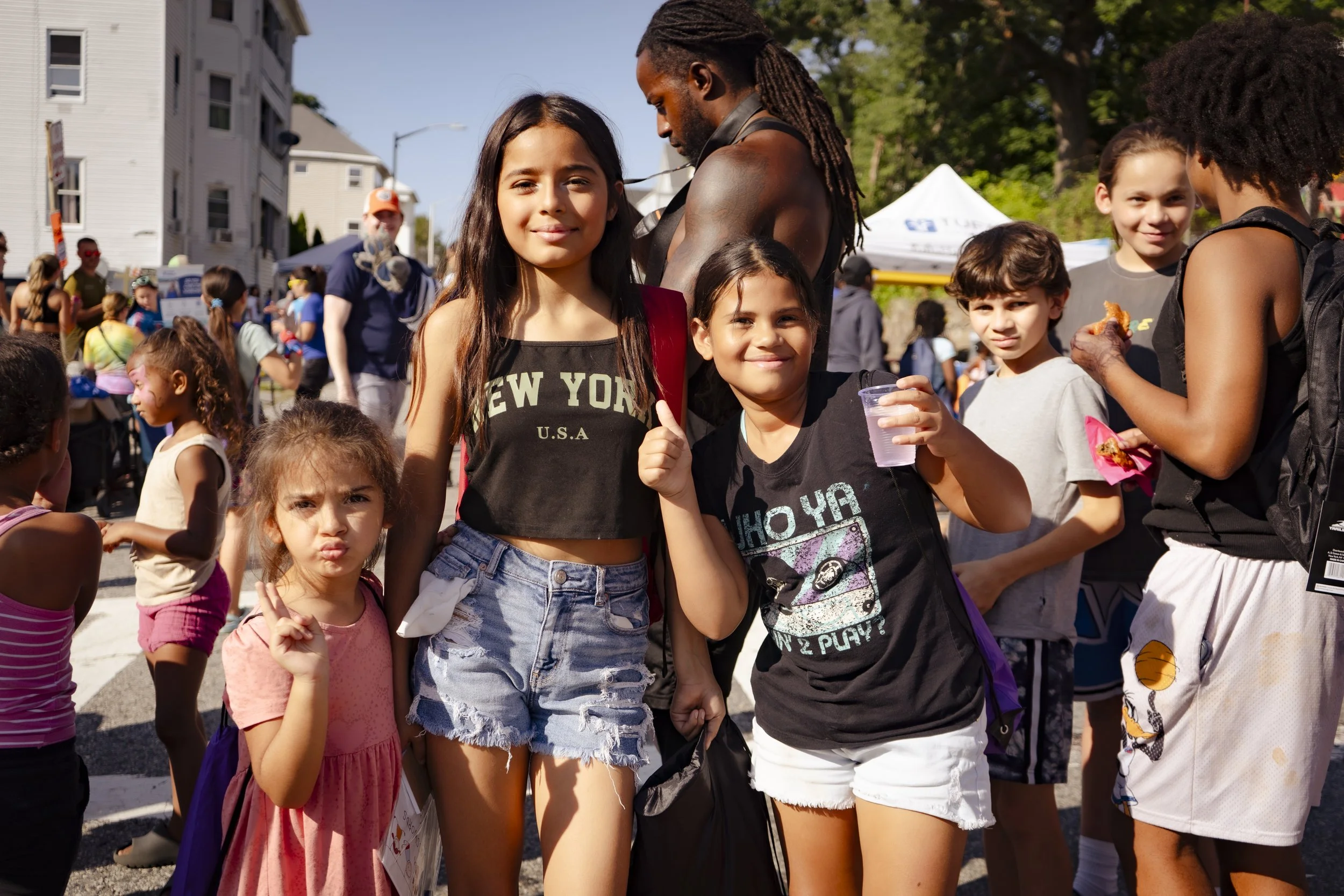 Friendly House - Children and a man gathered outdoors at a community event, some children holding food or drinks, with tents and buildings in the background.
