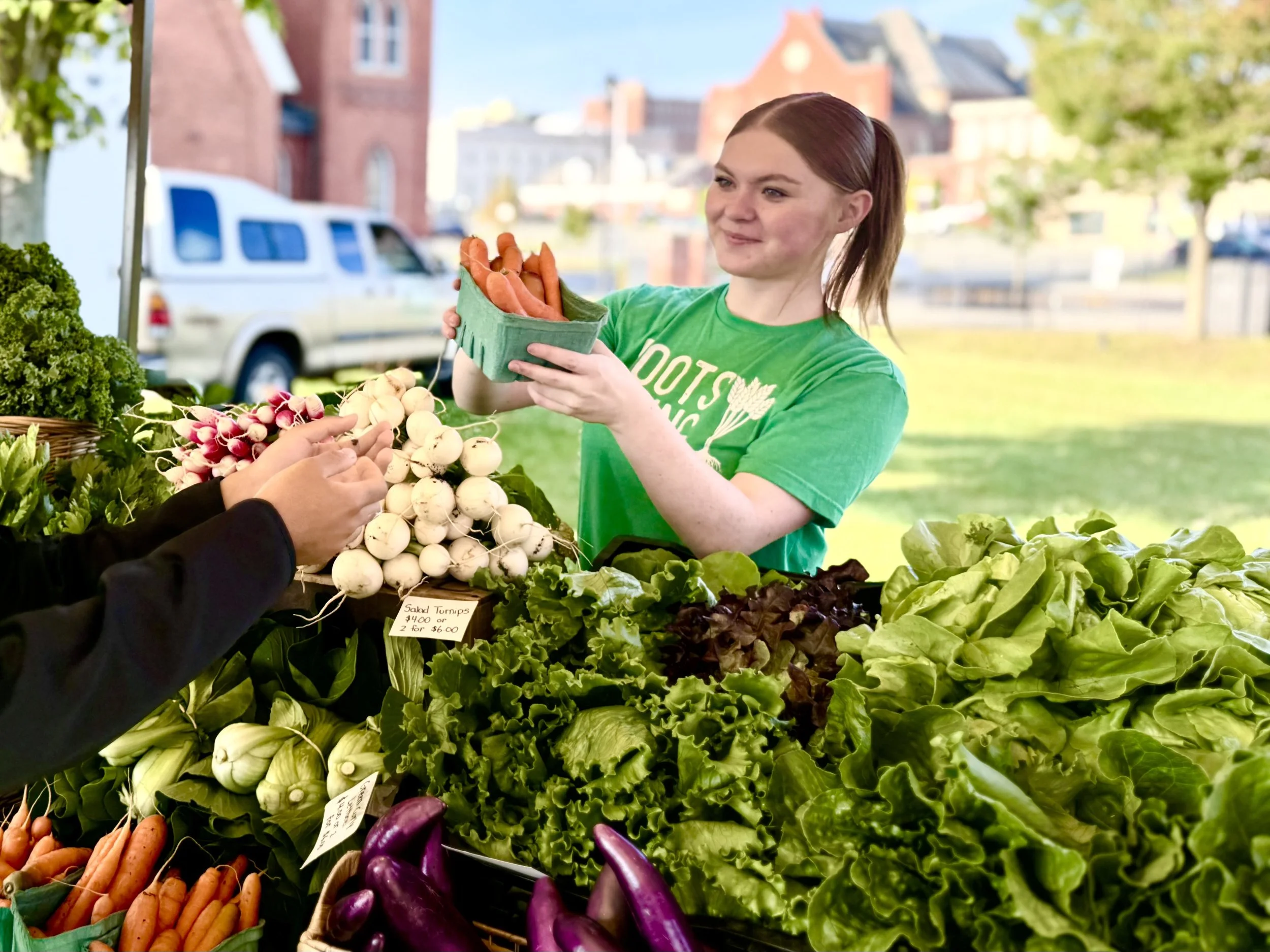 A woman with auburn hair wearing a green t-shirt is at a farmer's market stall with fresh vegetables. She is holding a green container of carrots and appears to be purchasing produce from a person with visible hands. The stall has various vegetables such as radishes, leafy greens, eggplants, and carrots, with a background of buildings and trees.