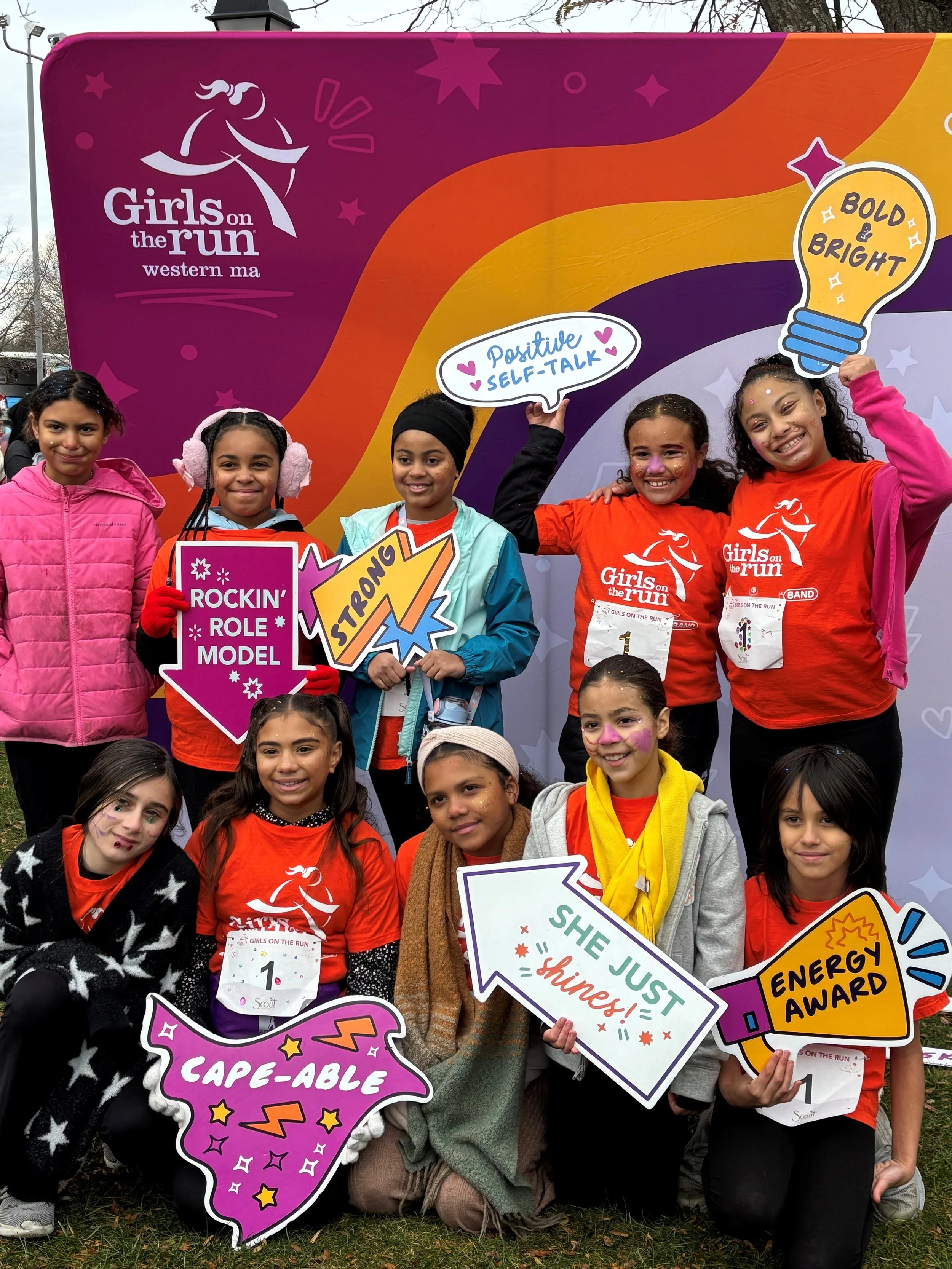 A group of young girls participating in the Girls on the Run event, holding colorful signs and wearing medals, standing in front of a vibrant backdrop. The backdrop features a rainbow, stars, and motivational phrases like "Positive Self-Talk," "Bold & Bright," and others, celebrating empowerment and confidence.