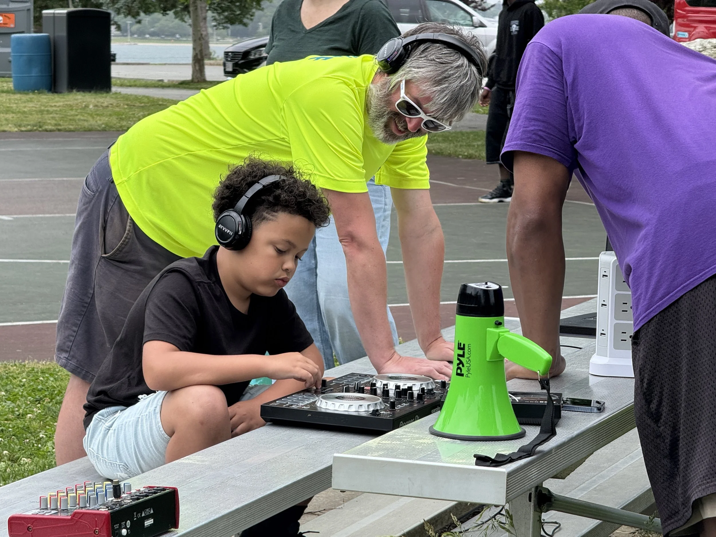 People gathered around outdoor table with DJ equipment, including a mixer, and drinks, engaging in an outdoor event with trees, a parking lot, and water in the background.