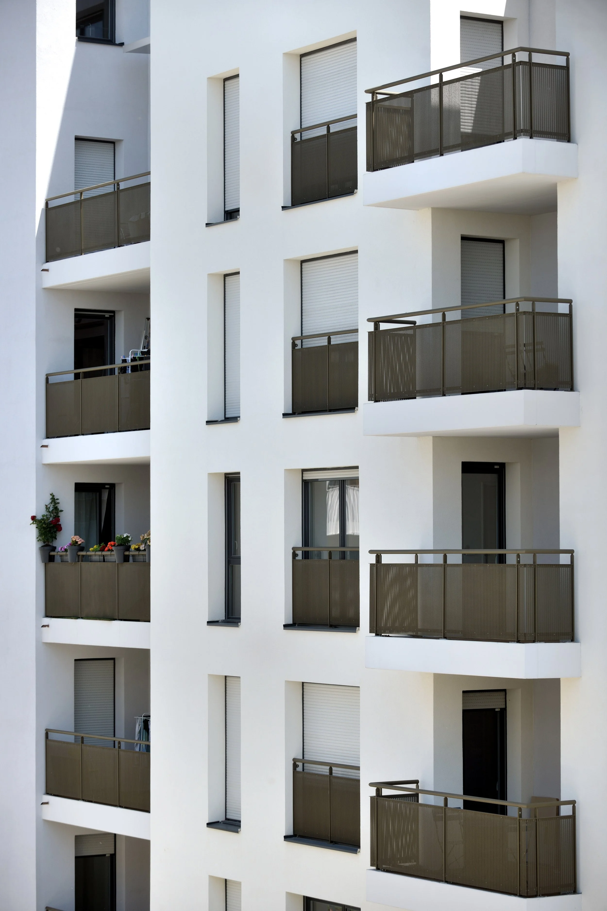 Immeuble résidentiel blanc avec plusieurs balcons équipés de garde-corps en métal brun, fenêtres à volets roulants fermés, quelques balcons avec des plantes. L'immeuble a un design moderne et épuré.