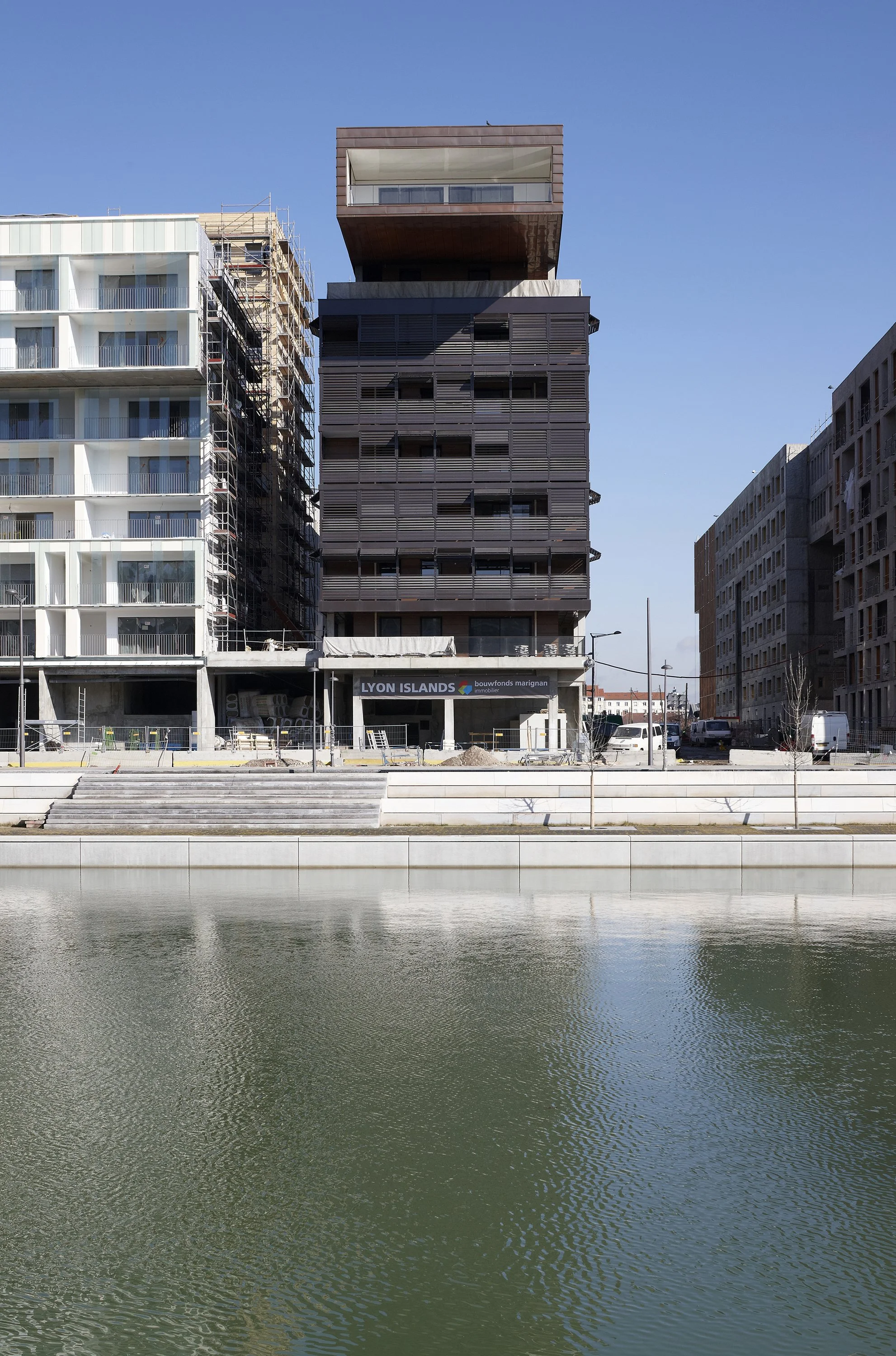 Un immeuble moderne en construction selon un style contemporain, situé au bord d'un plan d'eau, avec un ciel clair en arrière-plan.