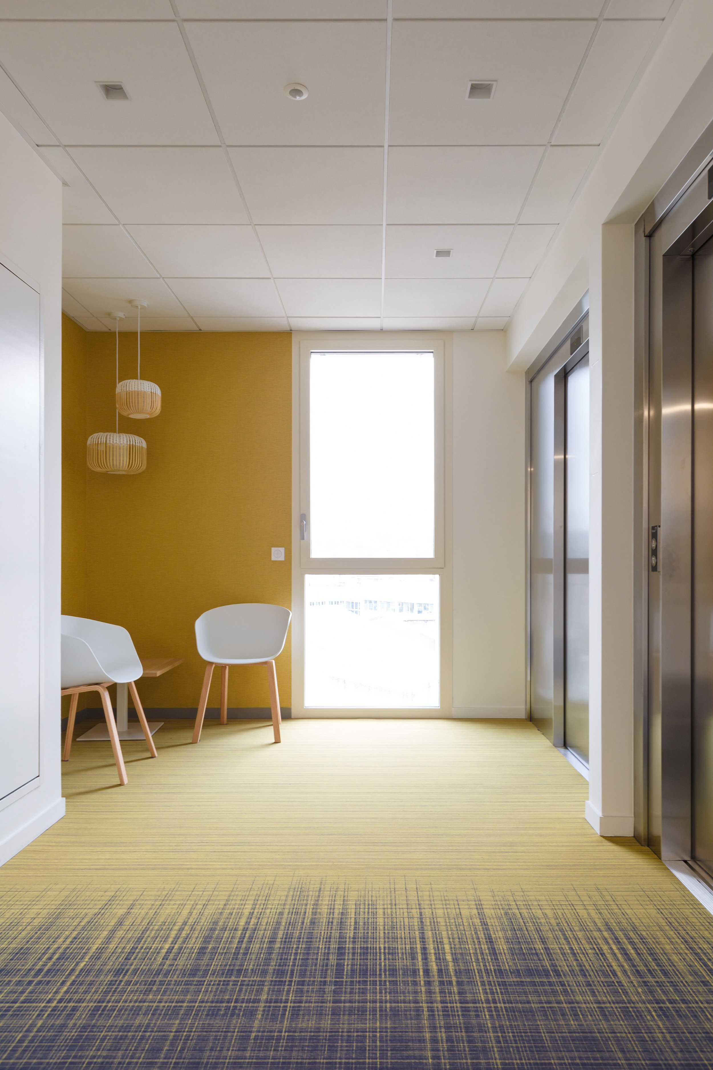 Un hall d'immeuble avec deux ascenseurs en acier, un mur jaune, une grande fenêtre, deux chaises blanches et deux lampes suspendues en bois.
