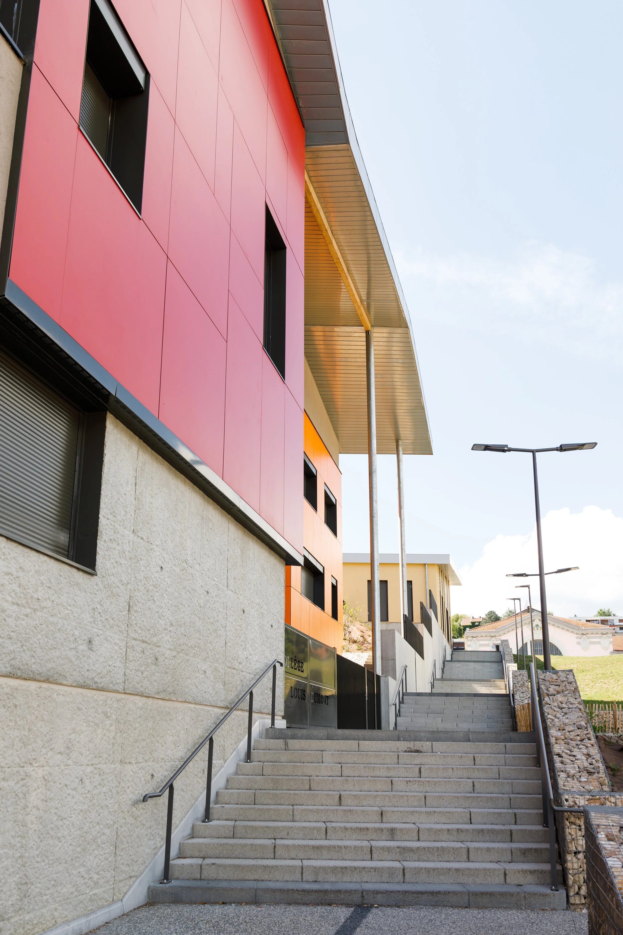 Escaliers extérieurs menant à un bâtiment avec façade colorée en rose et beige, lampadaires en alignement, et un ciel clair en arrière-plan.