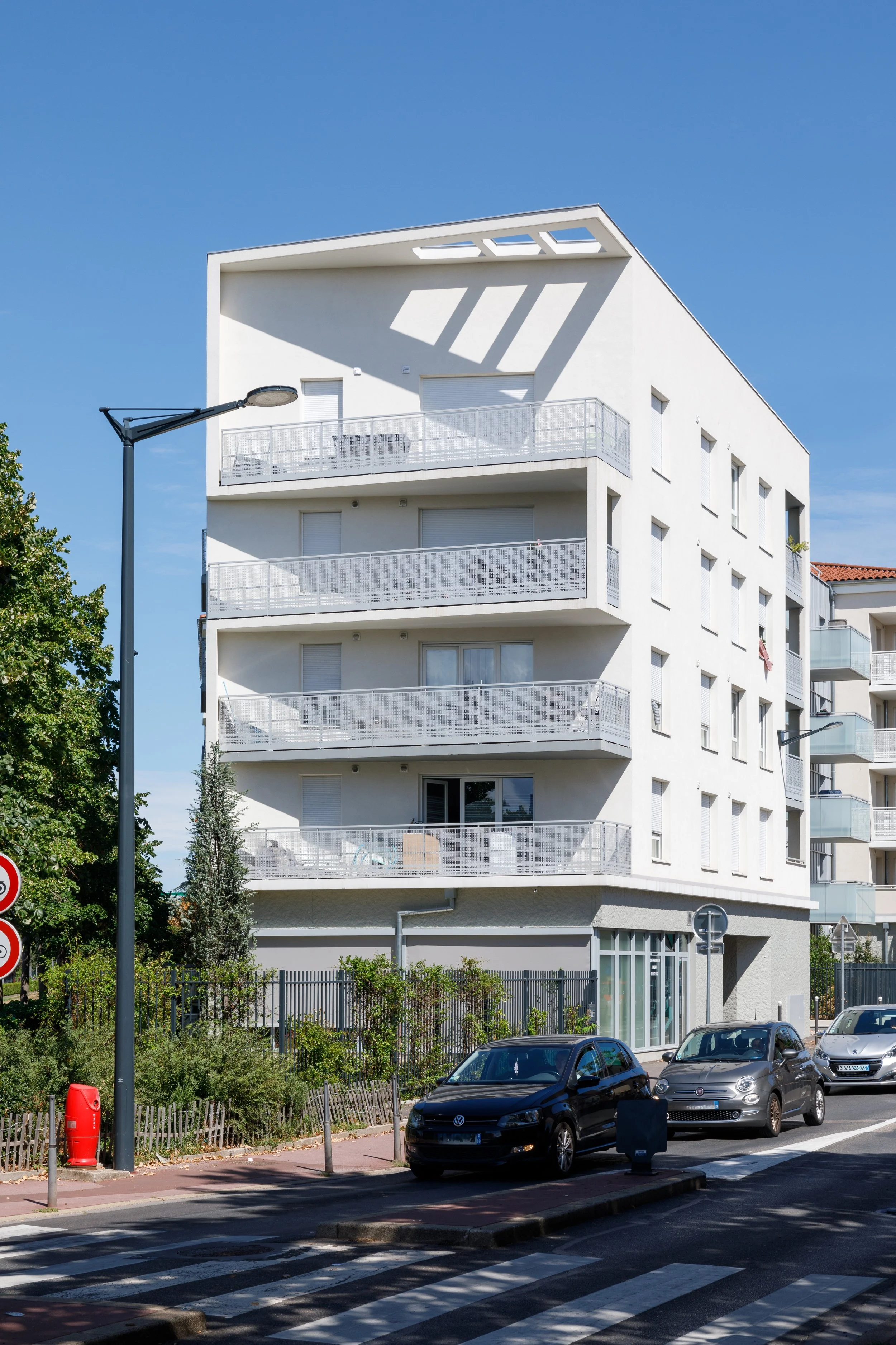 Une façade d'immeuble résidentiel blanc avec plusieurs balcons en métal, un lampadaire, et des voitures garées devant, sous un ciel bleu clair.